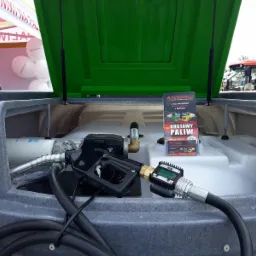 Mobilny dystrybutor paliwa z otwartą zieloną pokrywą, widoczny pistolet dystrybucyjny z elektronicznym licznikiem, butelki z płynem na obudowie, w tle ulotka reklamowa dostawcy paliw i balony...