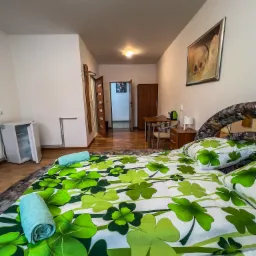 Pokój z łóżkiem w czterolistną koniczynę, zrolowane ręczniki, mała lodówka, drewniany stół z krzesłami i szafa w tle.