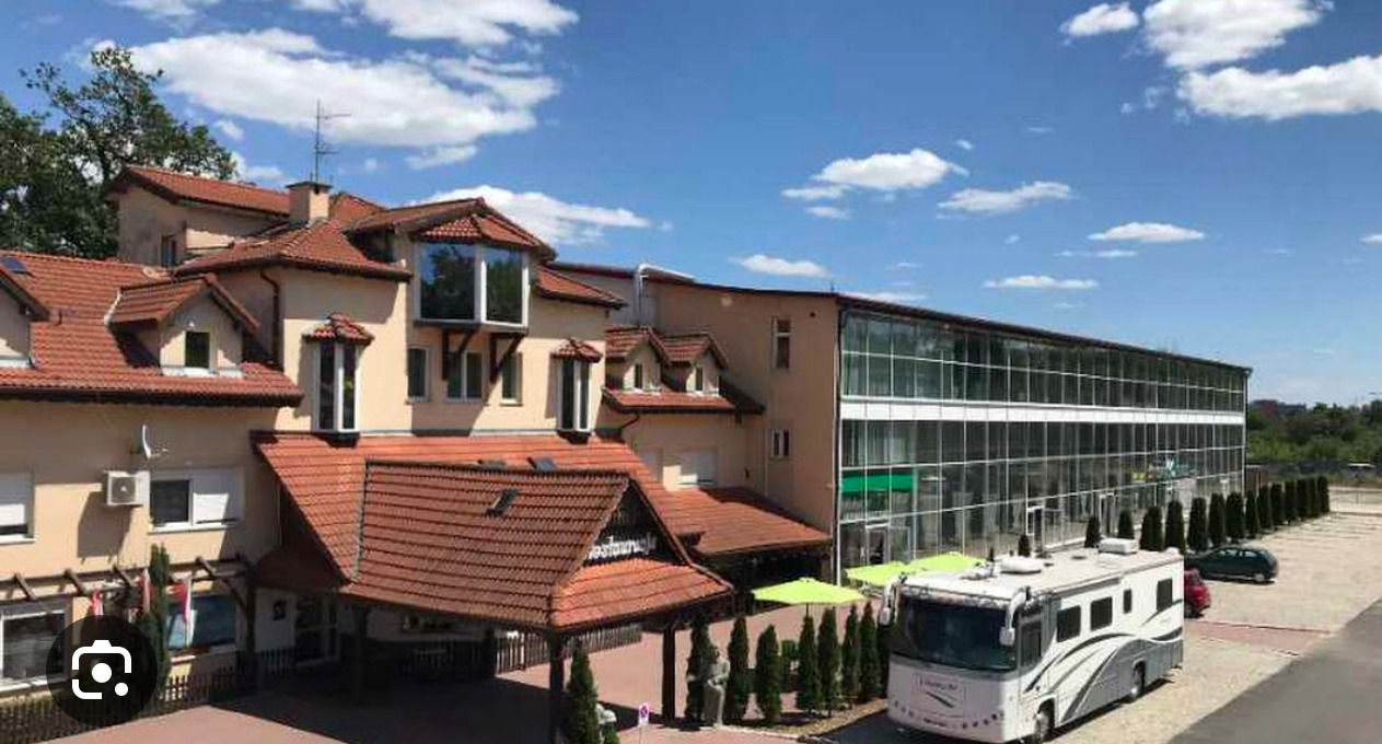 Kompleks budynków z parkingiem, widoczny budynek z czerwonym dachem i przeszklona hala, na parkingu zaparkowany kamper.