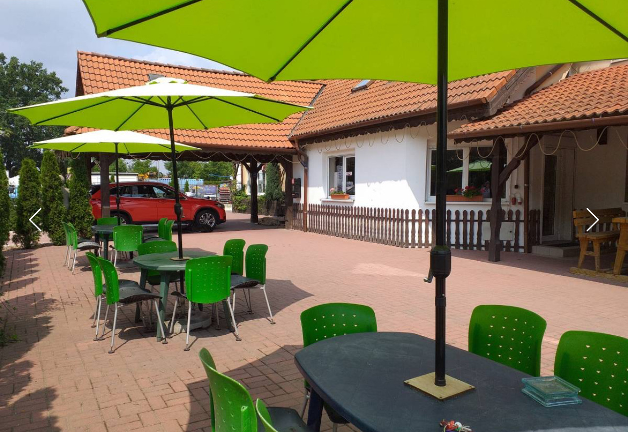 Zewnętrzne miejsce z zielonymi parasolami i meblami ogrodowymi, czerwony samochód zaparkowany w tle.