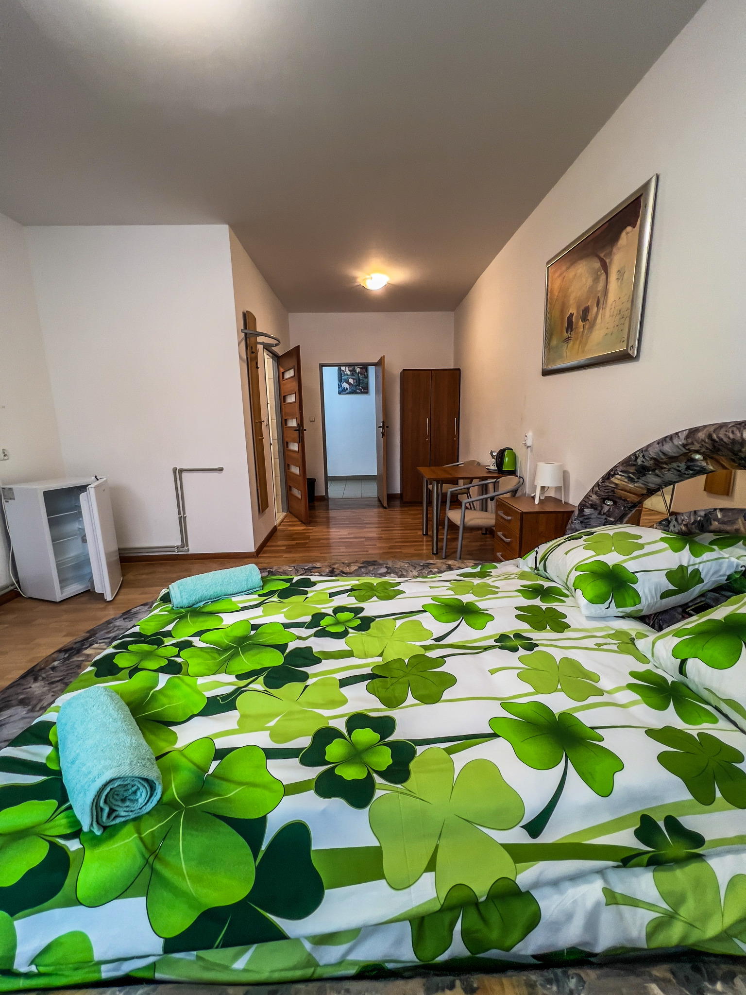 Pokój z łóżkiem w czterolistną koniczynę, zrolowane ręczniki, mała lodówka, drewniany stół z krzesłami i szafa w tle.