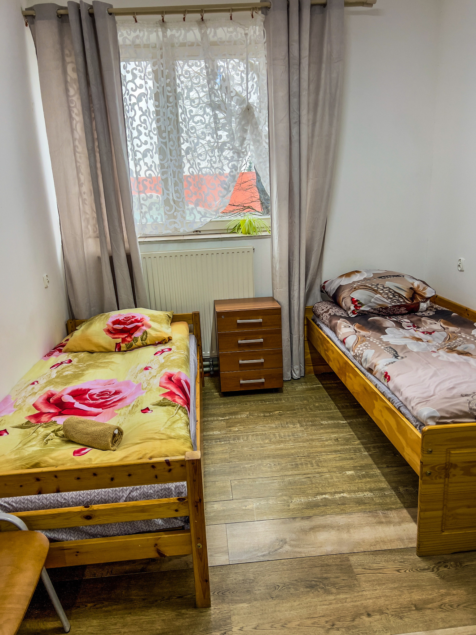 Pokój z dwoma pojedynczymi łóżkami o drewnianych ramach, pościel w kwiatowe wzory, drewniana szafka nocna z czterema szufladami, okno z firanką i zasłonami, drewniana podłoga.