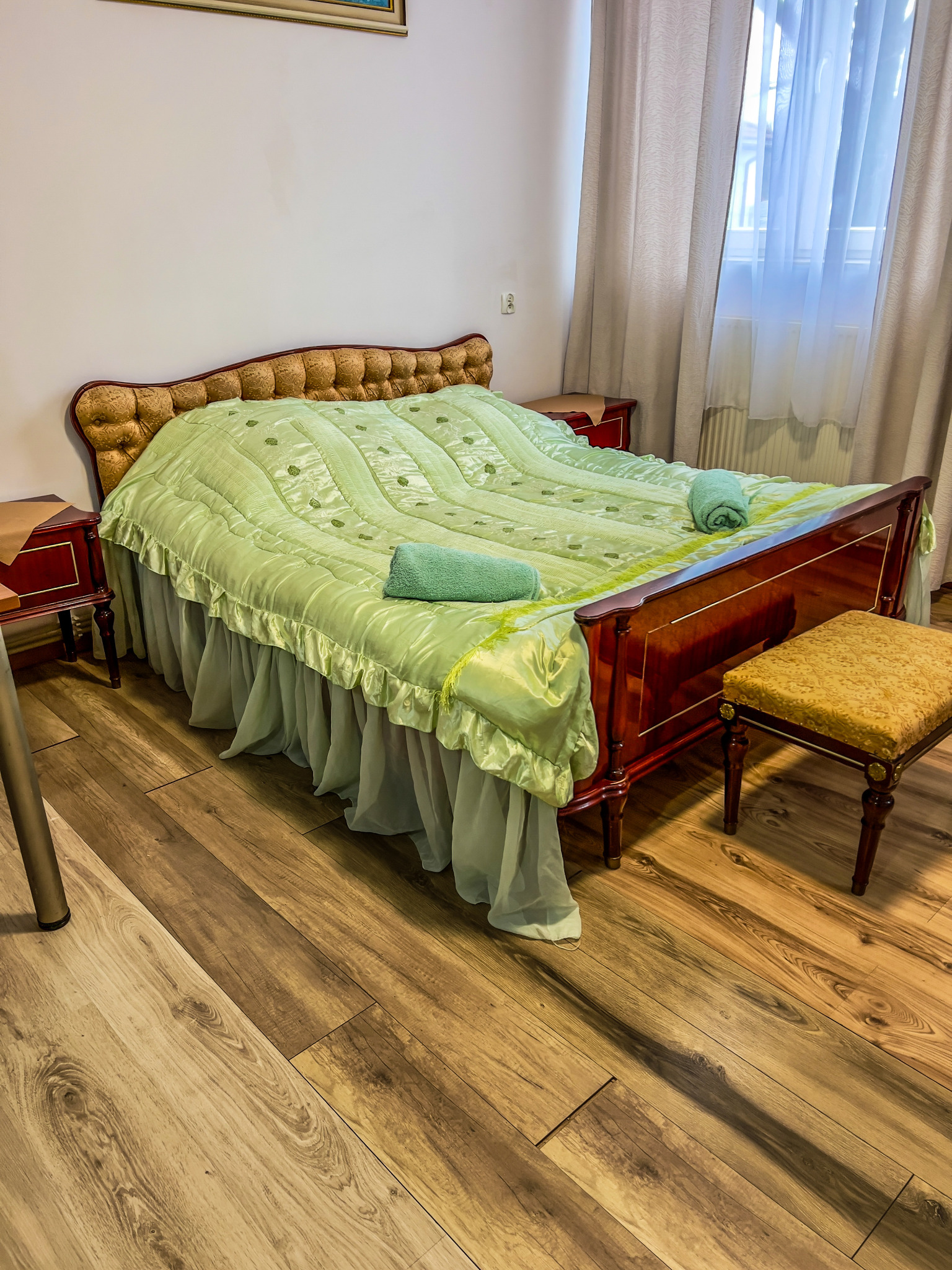 Pokój z łóżkiem o drewnianej ramie, nakrytym zieloną satynową narzutą z falbanami i dwoma zwiniętymi ręcznikami, obok stolik nocny i tapicerowany stołek na drewnianej podłodze.
