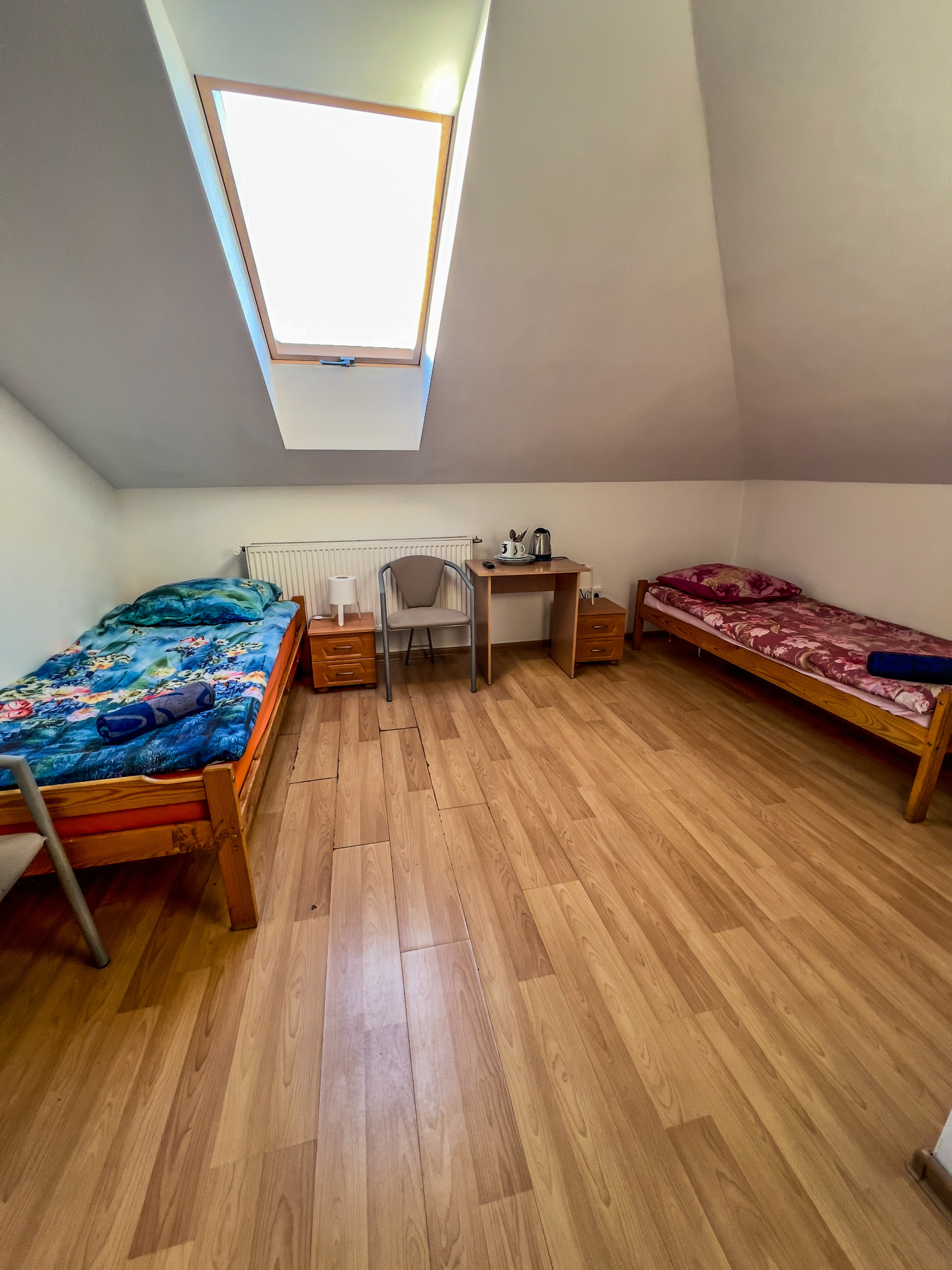 Pokój z dwoma łóżkami jednoosobowymi, drewnianą podłogą i oknem dachowym, wyposażony w stolik z czajnikiem i filiżankami.