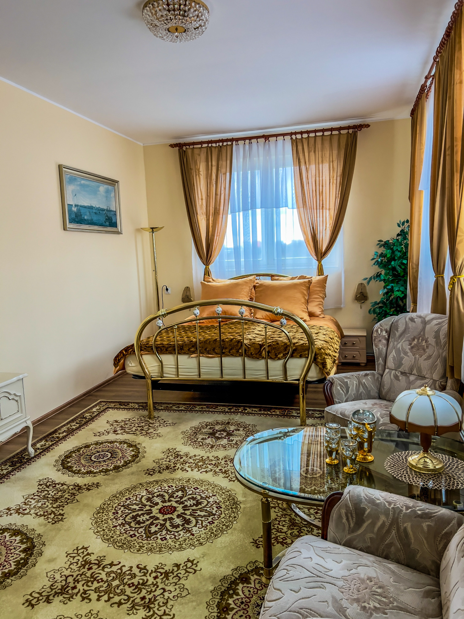 Elegancki pokój z łóżkiem o złotej ramie, zasłony w odcieniach złota, wzorzysty dywan i szklany stolik kawowy z dekoracyjnymi kieliszkami.