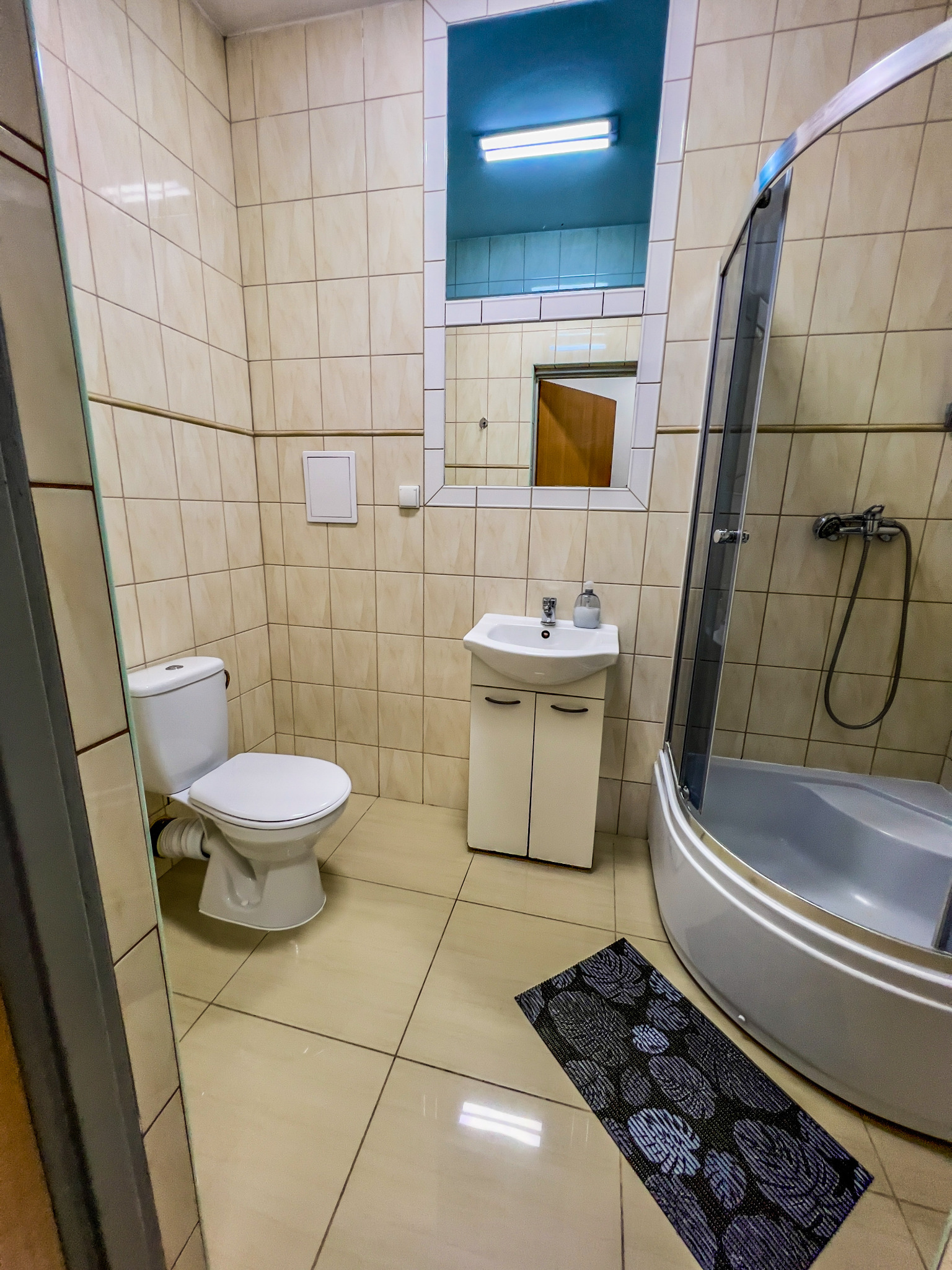 Wnętrze łazienki z kabiną prysznicową, umywalką z szafką, toaletą i czarnym dywanikiem na beżowych płytkach podłogowych, widok z perspektywy drzwi.