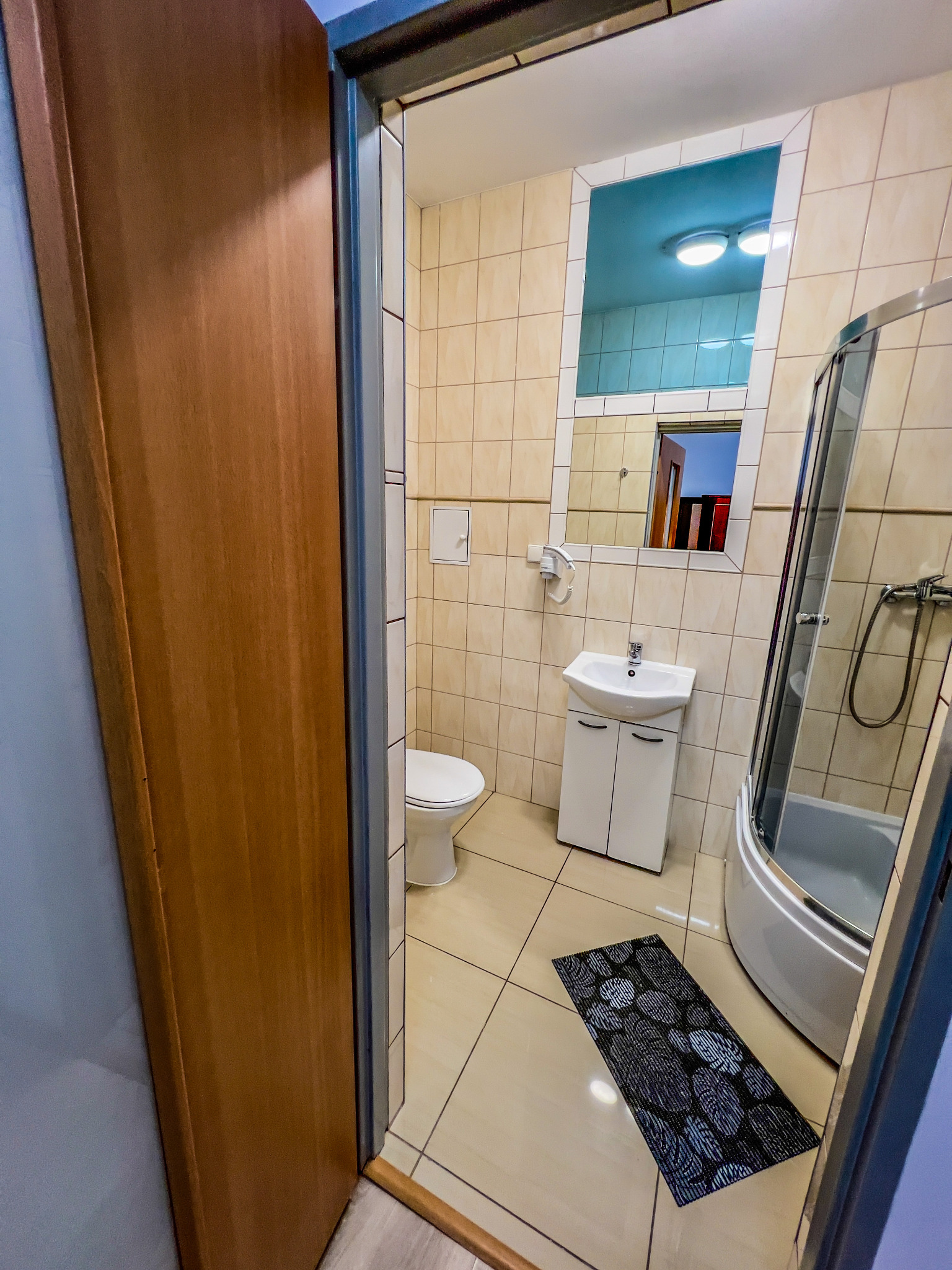 Wnętrze łazienki z kabiną prysznicową, umywalką z szafką i toaletą we Wrocławiu. Widoczny wzorzysty dywanik podłogowy i lustro.