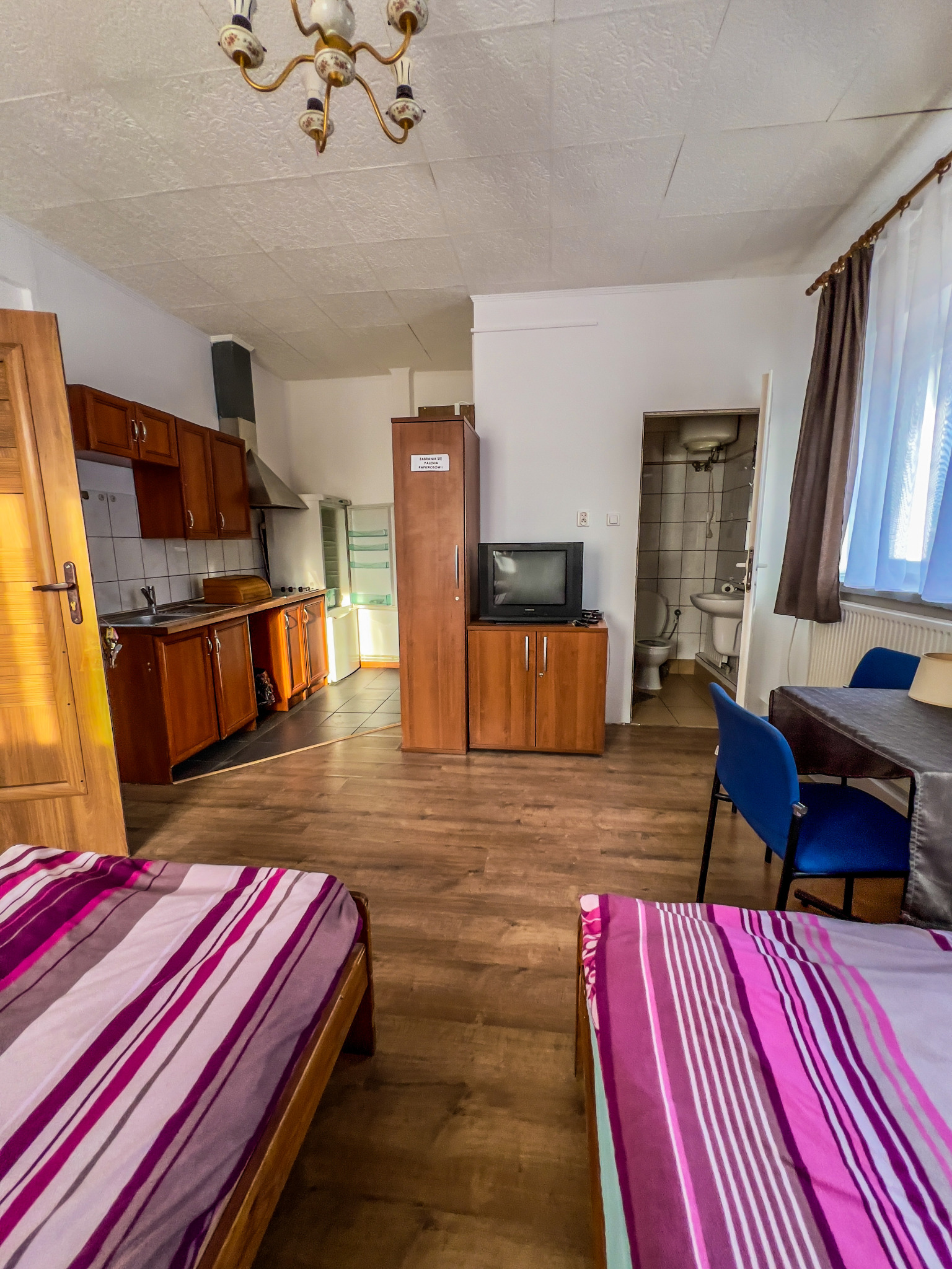 Widok wnętrza pokoju z aneksem kuchennym i łazienką, wyposażonego w drewniane meble, dwa łóżka z fioletowo-białą pościelą w paski oraz stół z niebieskimi krzesłami.