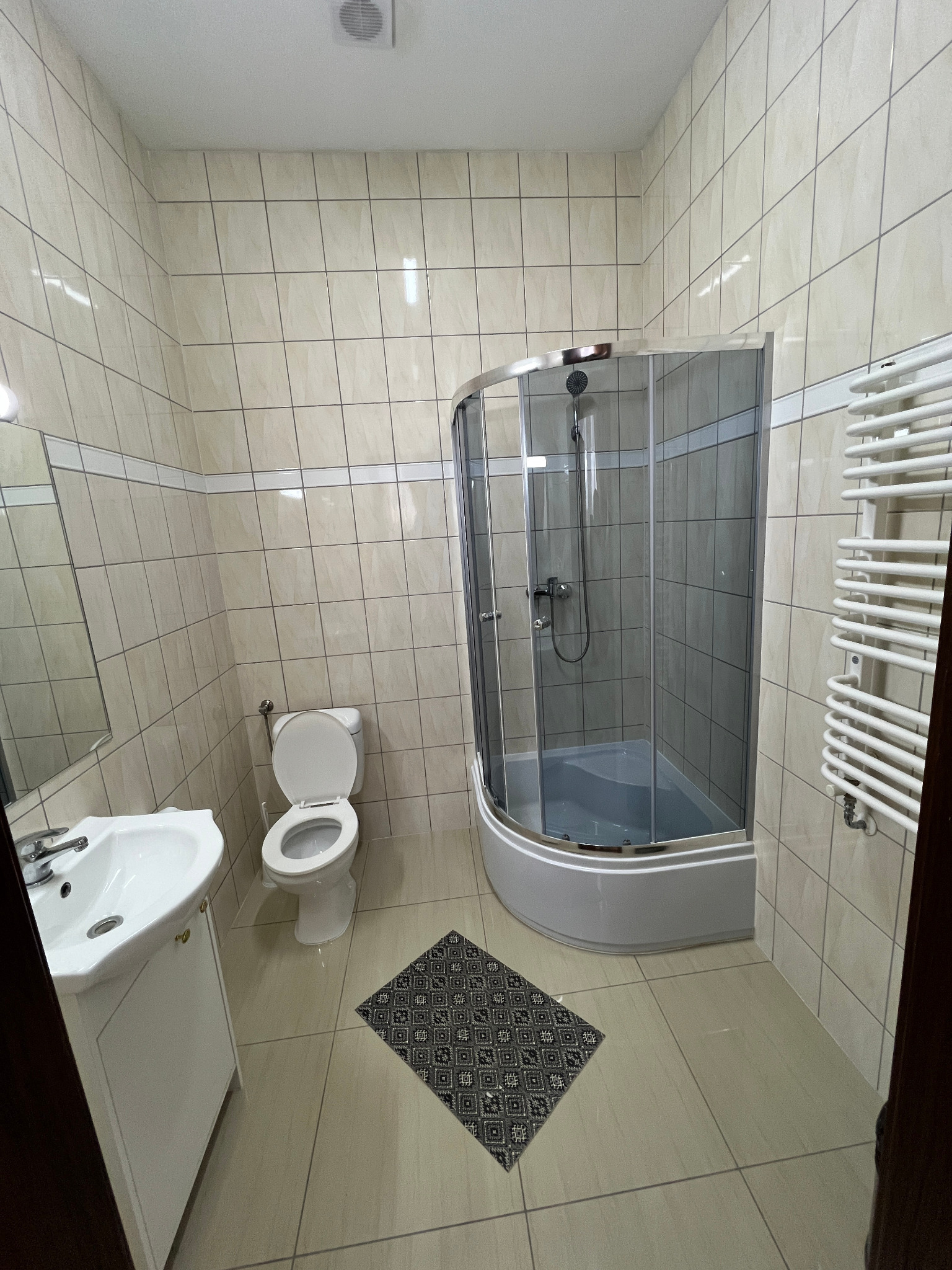 Jasna łazienka z beżowymi płytkami, kabiną prysznicową z zaokrąglonymi drzwiami, białą umywalką z szafką, toaletą z otwartą klapą i grzejnikiem drabinkowym na ścianie. Mały dywanik z geometrycznym...