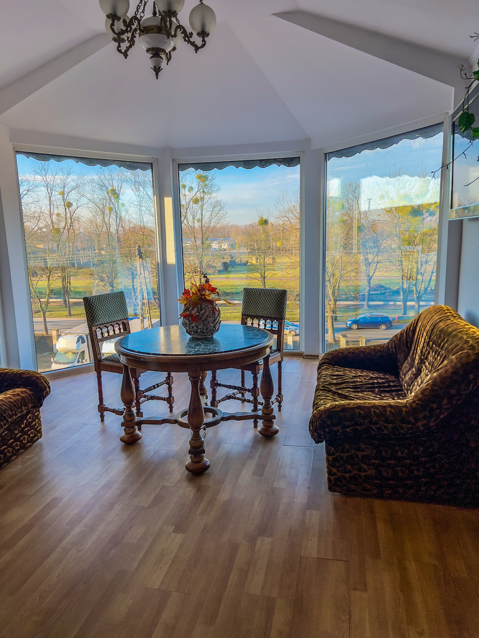 Jasne wnętrze z panoramicznym oknem, okrągłym stołem z dekoracją, dwoma krzesłami i wzorzystą sofą w odcieniach brązu.