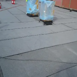 Płaski dach pokryty papą bitumiczną, widoczne zabezpieczone kominy i gaśnice w tle.