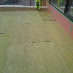 Płyty izolacyjne Rockwool ułożone na poddaszu, widoczna ściana z oknem i fragment odblaskowej kamizelki w rogu