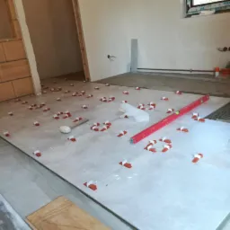 Układanie jasnych płytek podłogowych z użyciem poziomicy i klipsów dystansowych w remontowanym pomieszczeniu. Widoczny gumowy młotek i fragment ściany z cegły.