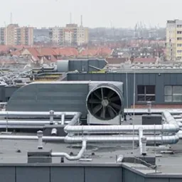 Widok z góry na system wentylacyjny z dużym wentylatorem na dachu budynku, z widoczną panoramą miasta w tle i dźwigami portowymi w oddali.