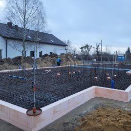 Bodnenplatte Sp. z o.o. - Zbrojenie fundamentów z użyciem bloczków styropianowych, widoczna siatka z prętów stalowych, wystające pionowe pręty, w tle budynek mieszkalny i sterta ziemi.