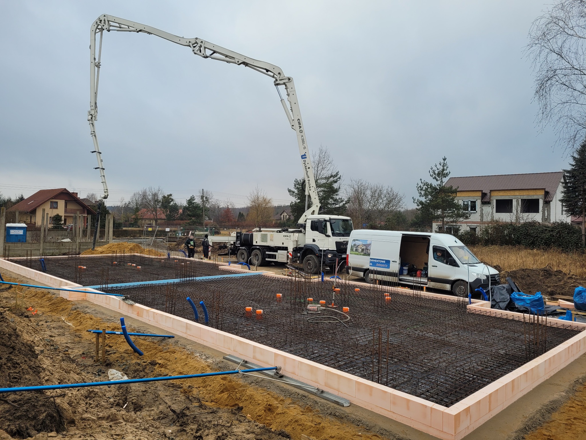 Zbrojenie fundamentów z ułożonymi rurami, otoczone termoizolacją, z pompą do betonu i ekipą budowlaną w tle.