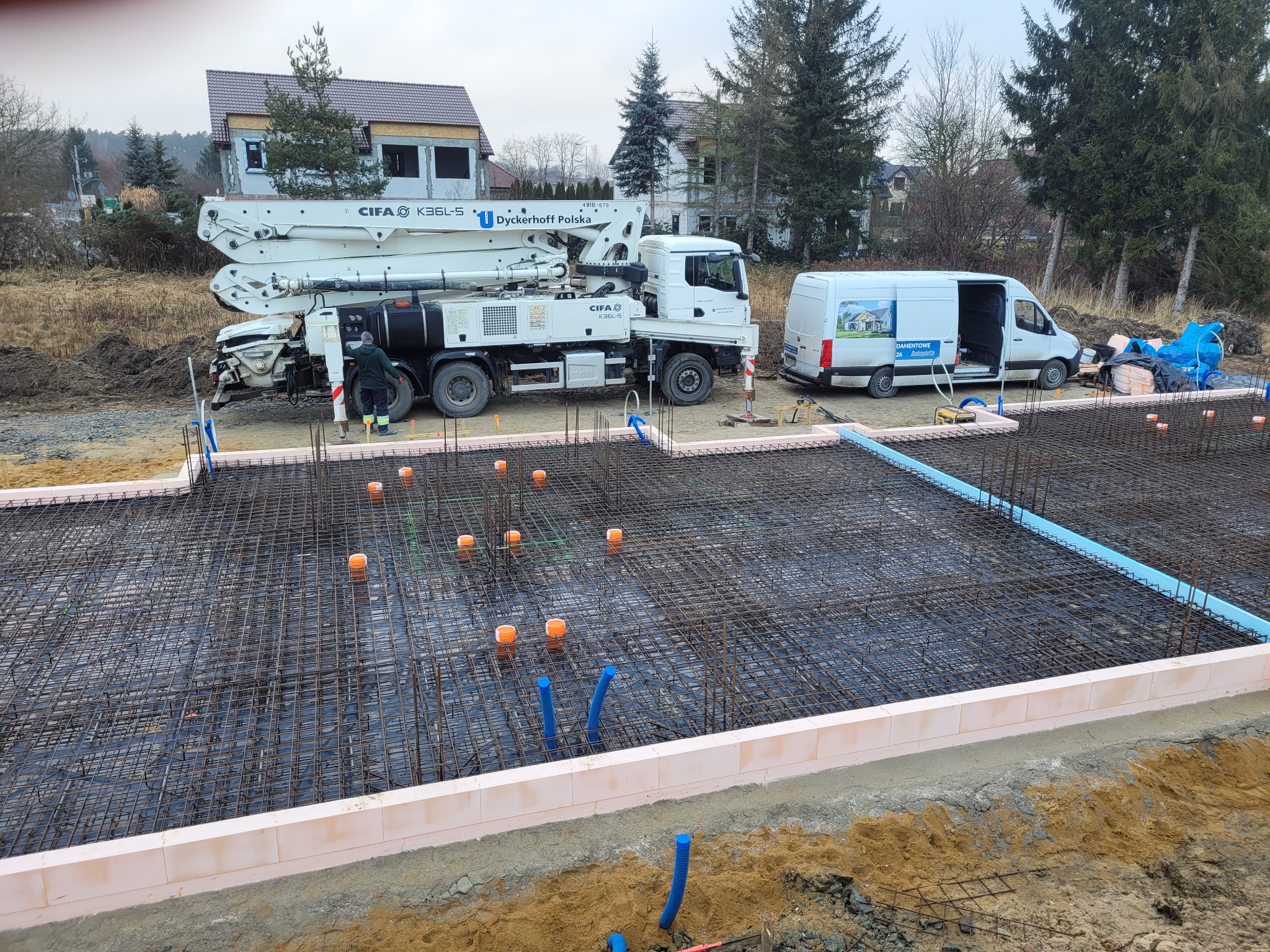 Zbrojenie fundamentu w szalunku styropianowym z widocznymi pionowymi rurami instalacyjnymi, pompą do betonu CIFA K36L-5 i samochodem dostawczym z reklamą firmy w tle.