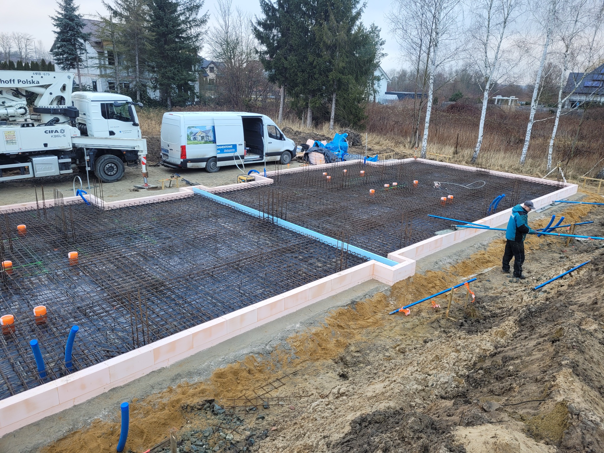 Zbrojenie fundamentu z siatki stalowej, obłożone termoizolacją z jasnoróżowego styroduru, z widocznymi niebieskimi rurami instalacyjnymi i pracownikiem w niebieskiej kurtce, na tle betonomieszarki...