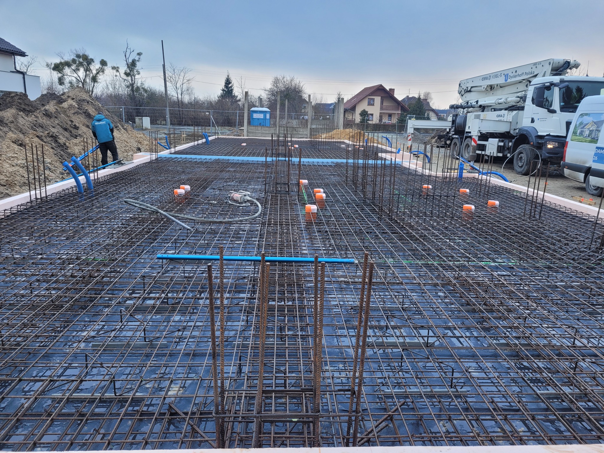 Zbrojenie fundamentu domu jednorodzinnego z widocznymi niebieskimi rurami instalacji i pompą do betonu w tle.