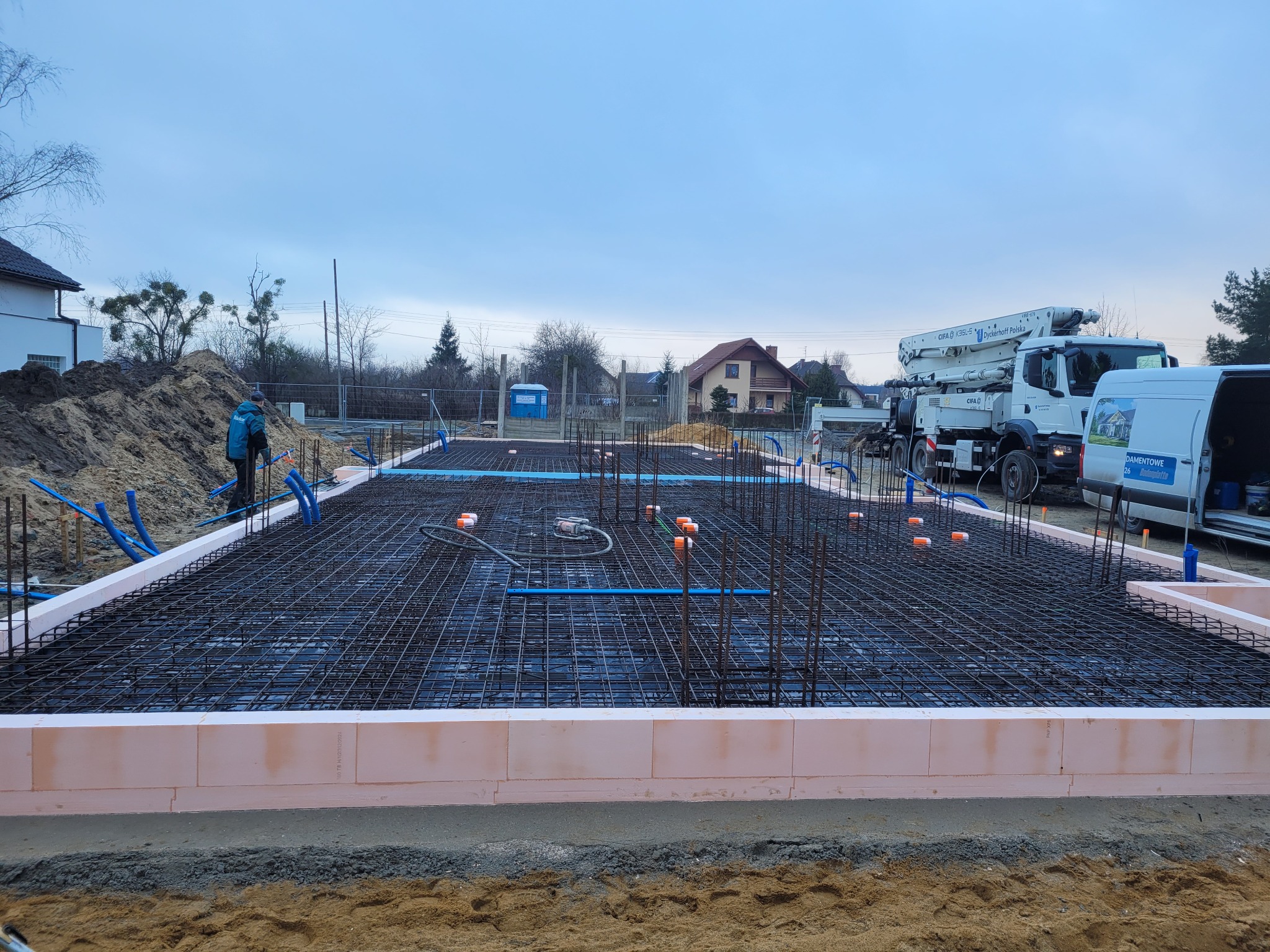 Przygotowany zbrojony szalunek z izolacją termiczną pod płytę fundamentową z widocznymi pionowymi prętami, rurami instalacyjnymi i pompą do betonu w tle.