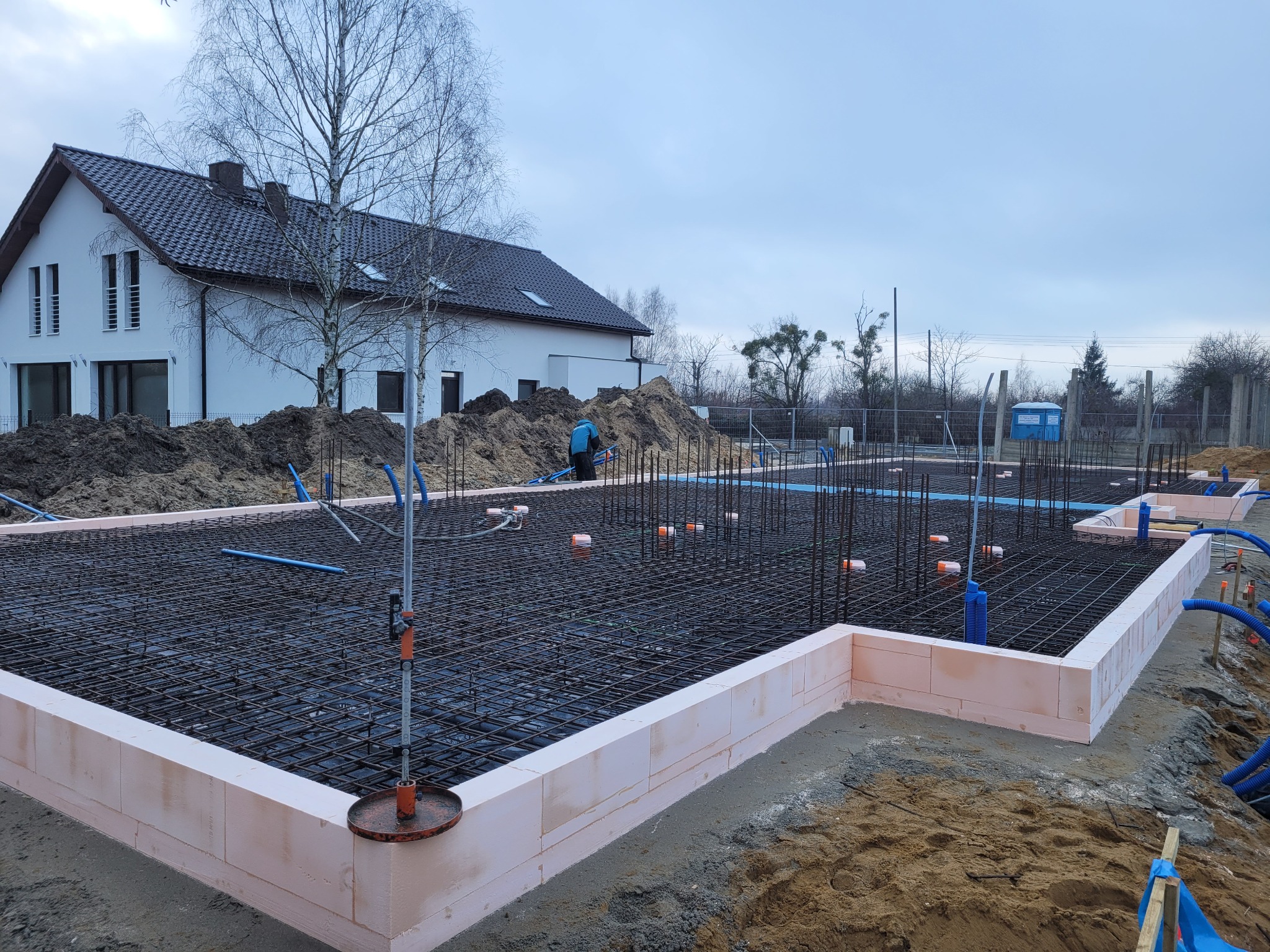 Zbrojenie fundamentów z użyciem bloczków styropianowych, widoczna siatka z prętów stalowych, wystające pionowe pręty, w tle budynek mieszkalny i sterta ziemi.