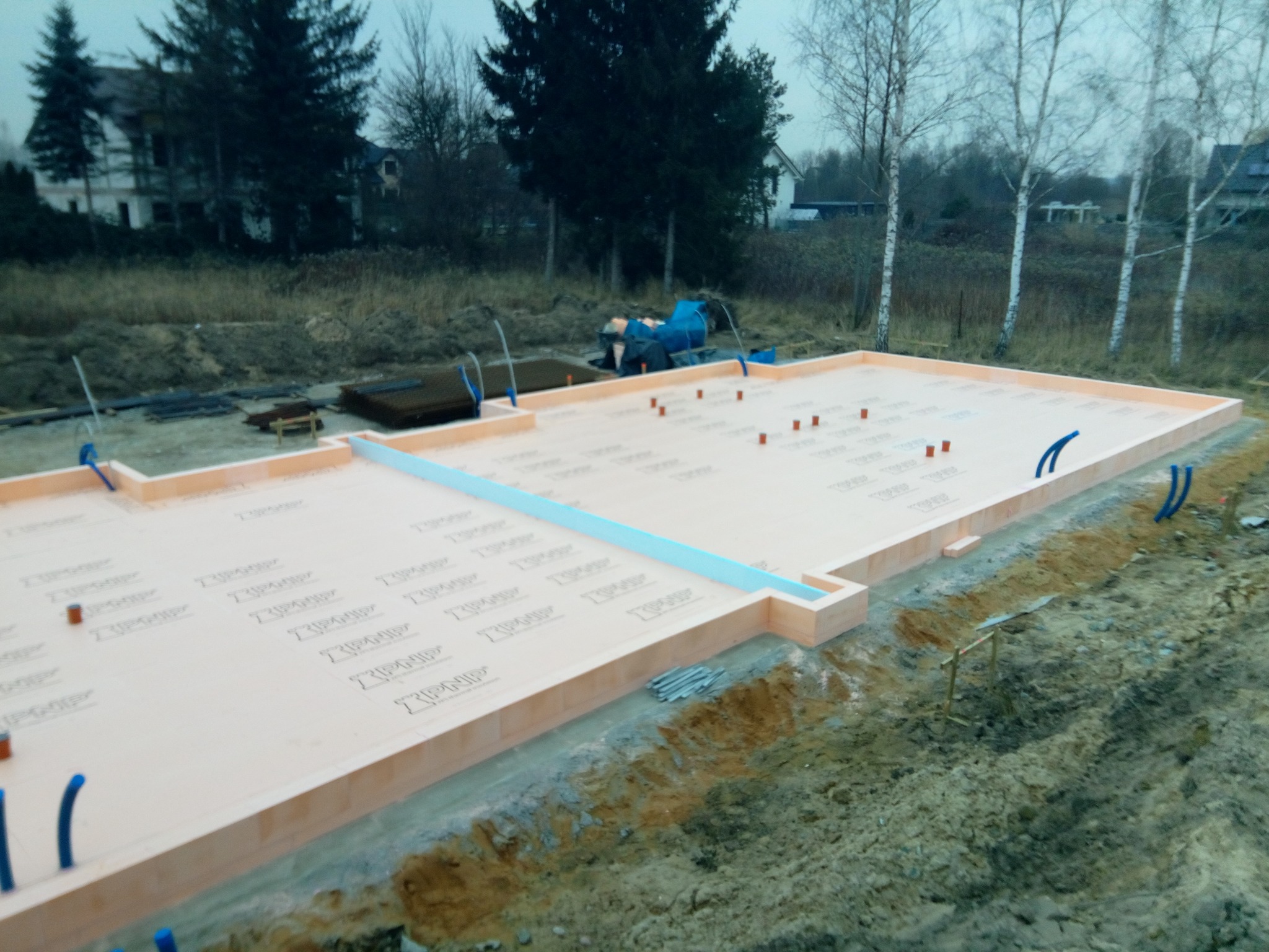 Przygotowany fundament z izolacją termiczną, widoczne wystające rurki instalacyjne, teren budowy z drzewami w tle.