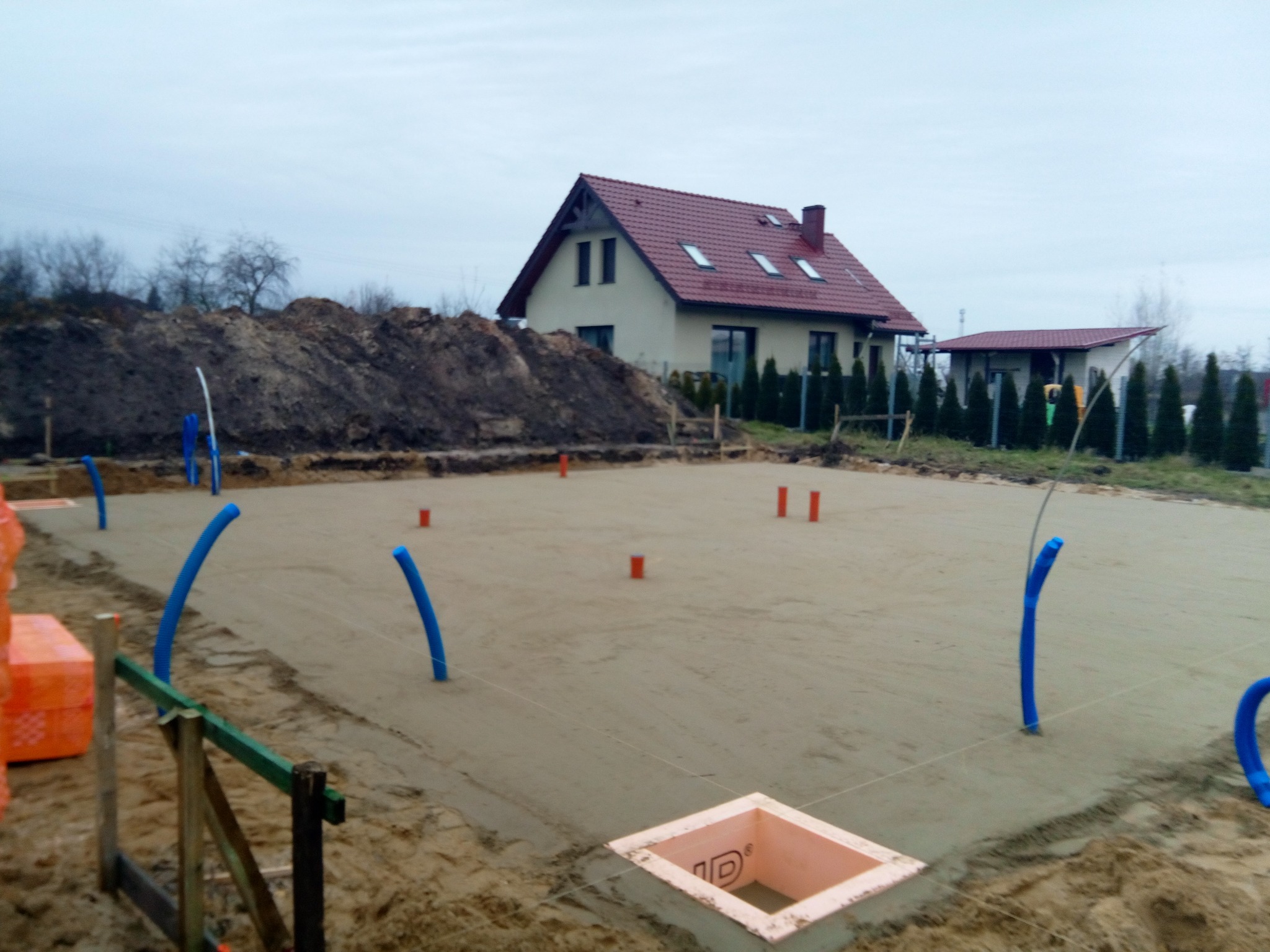 Przygotowany teren pod fundamenty w Trzebnicy z widocznymi instalacjami i wykopami pod ławy fundamentowe, w tle dom jednorodzinny z czerwoną dachówką.