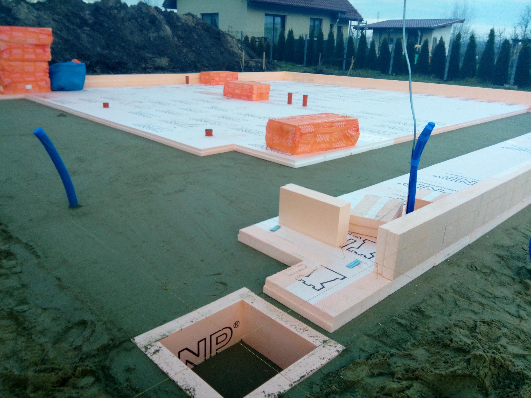 Budowa fundamentów: izolacja termiczna z płyt XPS, widoczne elementy systemu szalunkowego i instalacje w gruncie, w tle dom jednorodzinny.