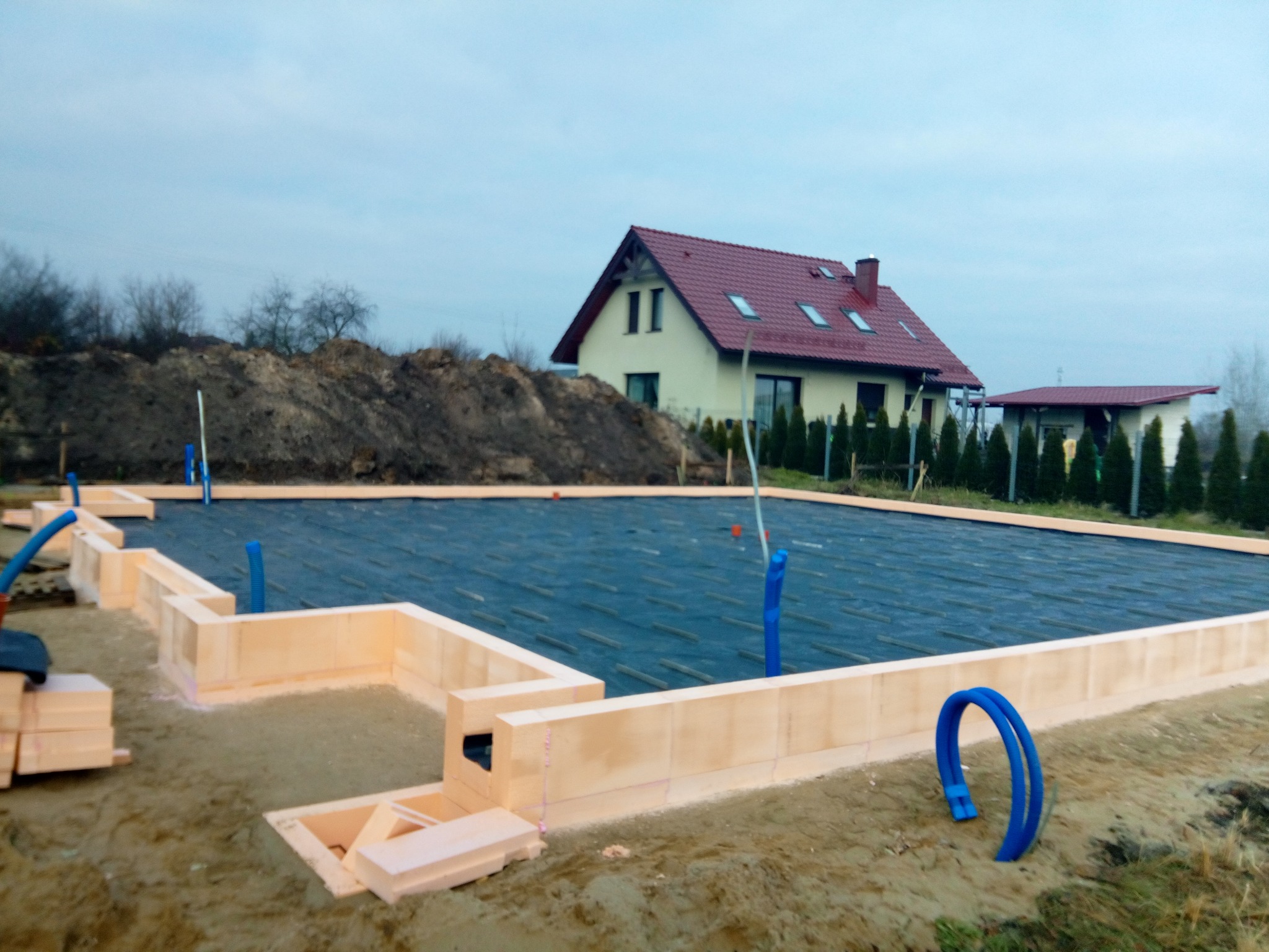 Budowa fundamentów domu: widok na formę z izolacji termicznej, czarną membranę hydroizolacyjną, wystające rury instalacyjne, w tle ziemia i budynek mieszkalny.