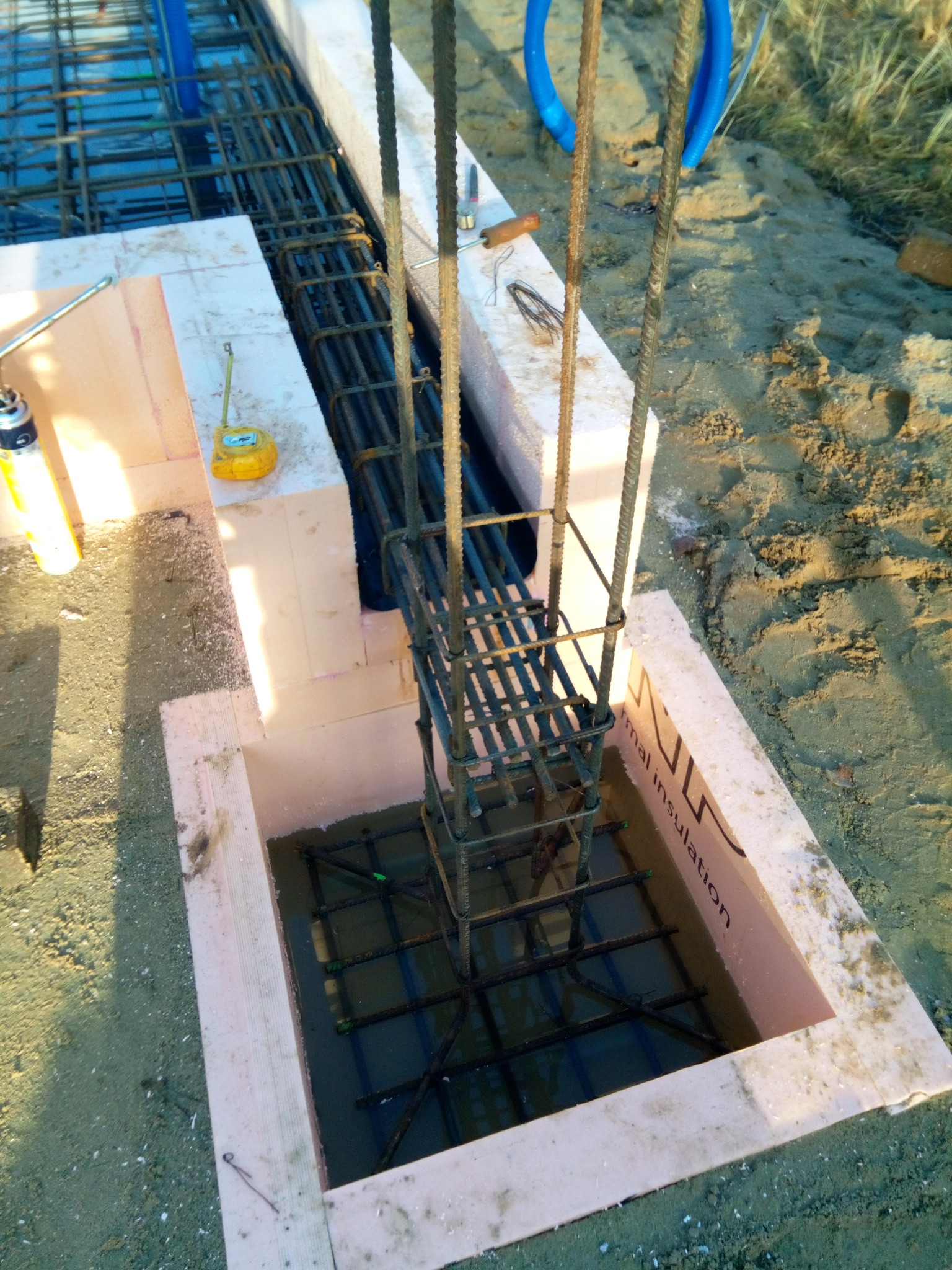 Zbrojenie pionowe i poziome w szalunku z termoizolacji, przygotowane do zalania betonem, widoczna żółta miarka zwijana i narzędzia budowlane w tle.