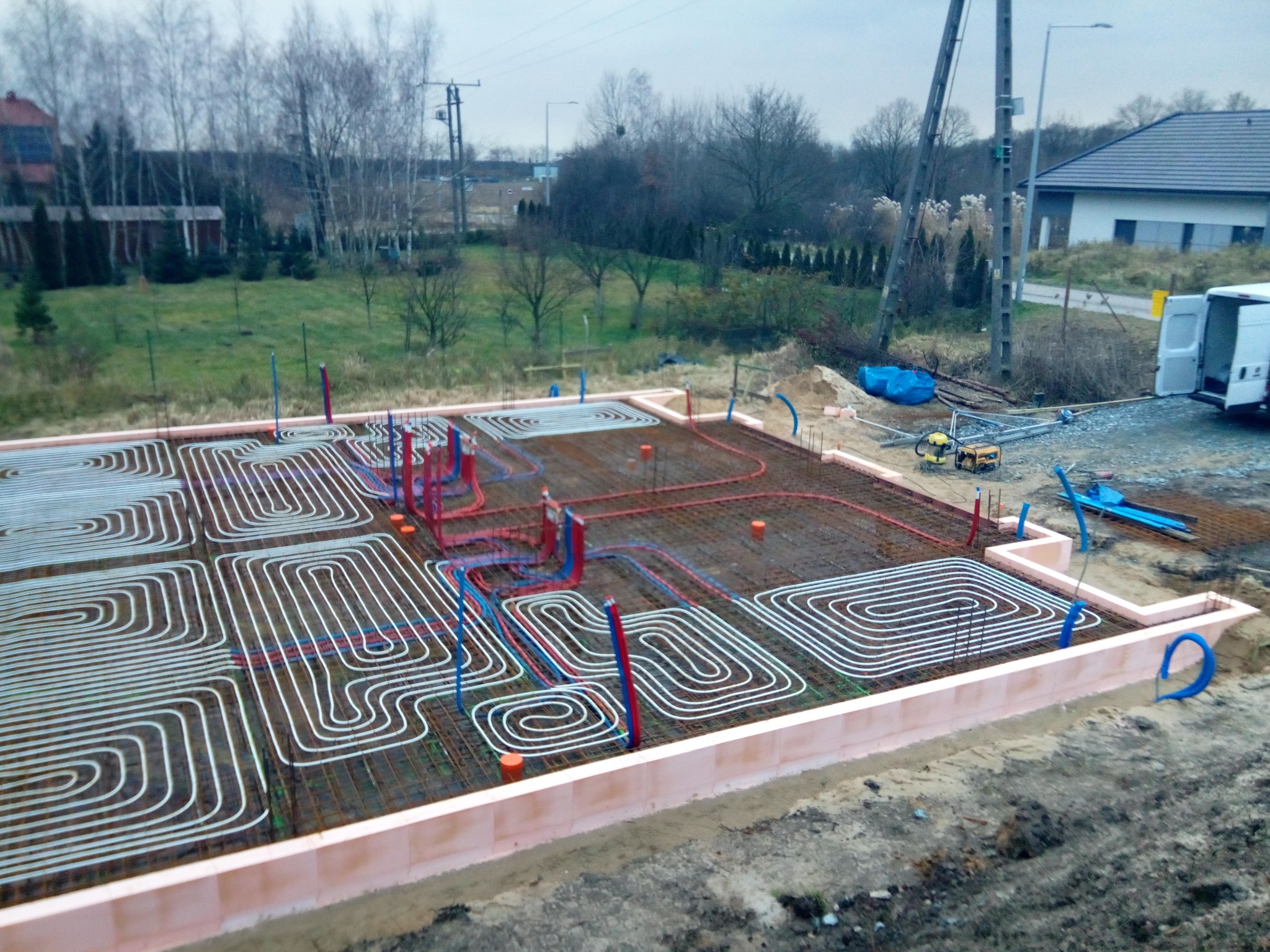 Przygotowanie fundamentów pod ogrzewanie podłogowe: rury grzewcze ułożone w spiralne wzory na siatce zbrojeniowej, otoczone izolacją termiczną, z widocznym sprzętem budowlanym w tle.