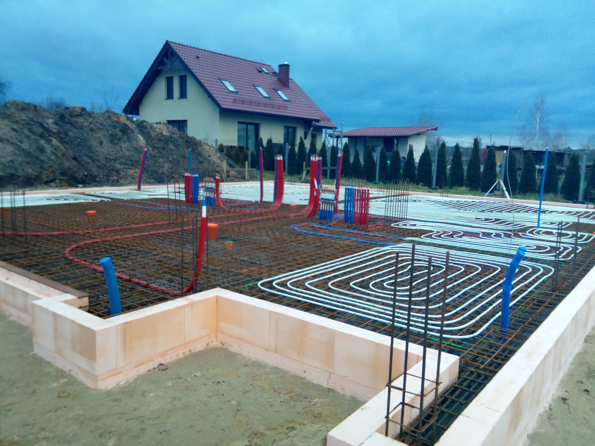 Fundamenty domu jednorodzinnego z widocznym systemem ogrzewania podłogowego, rury w kolorze białym układane na siatce zbrojeniowej, ściany fundamentowe z bloczków izolacyjnych, w tle dom w trakcie...