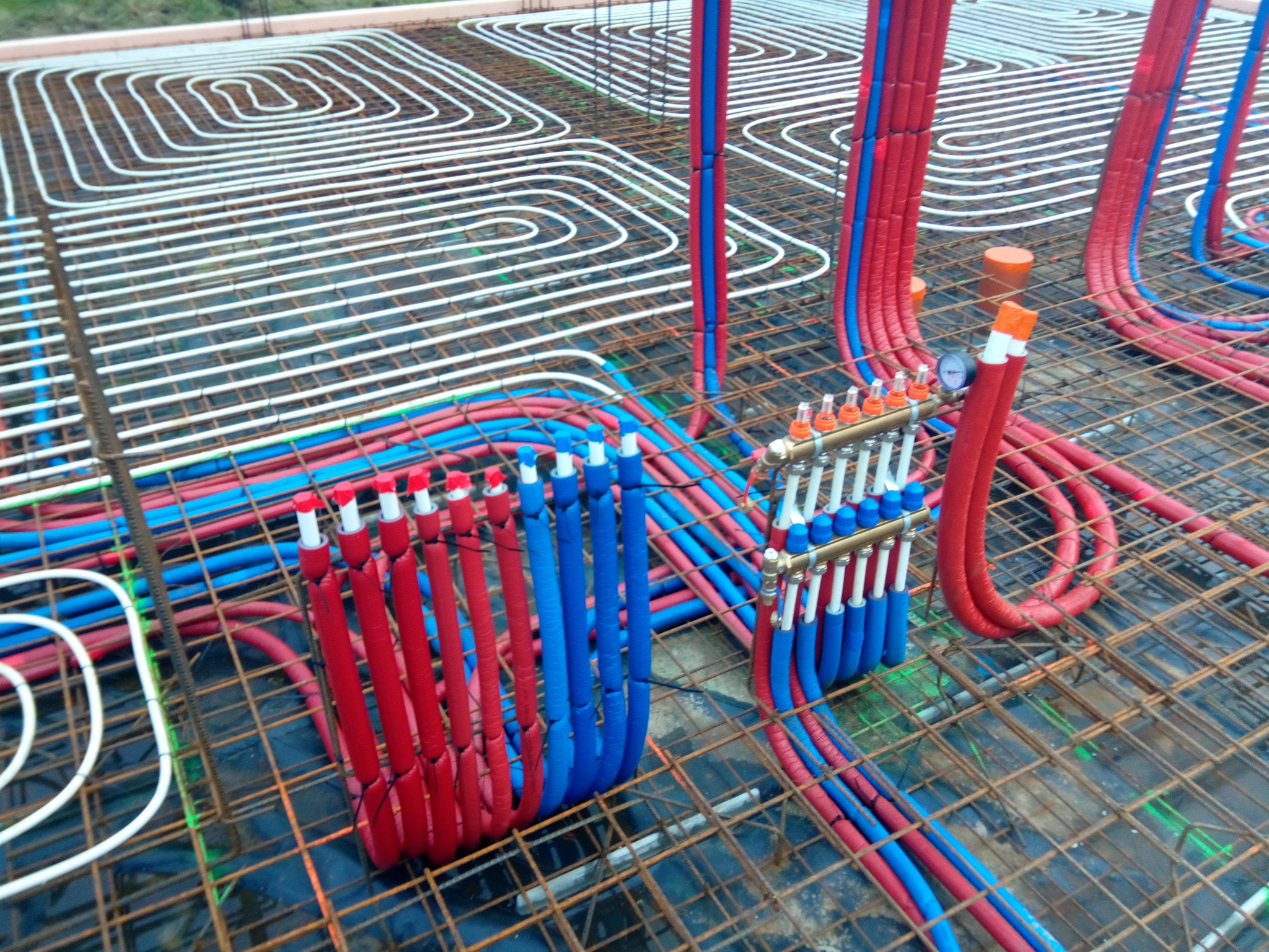 Rozłożone rury ogrzewania podłogowego w kolorach białym, czerwonym i niebieskim, zamocowane do metalowej siatki z rozdzielaczem, przygotowane do zalania betonem.