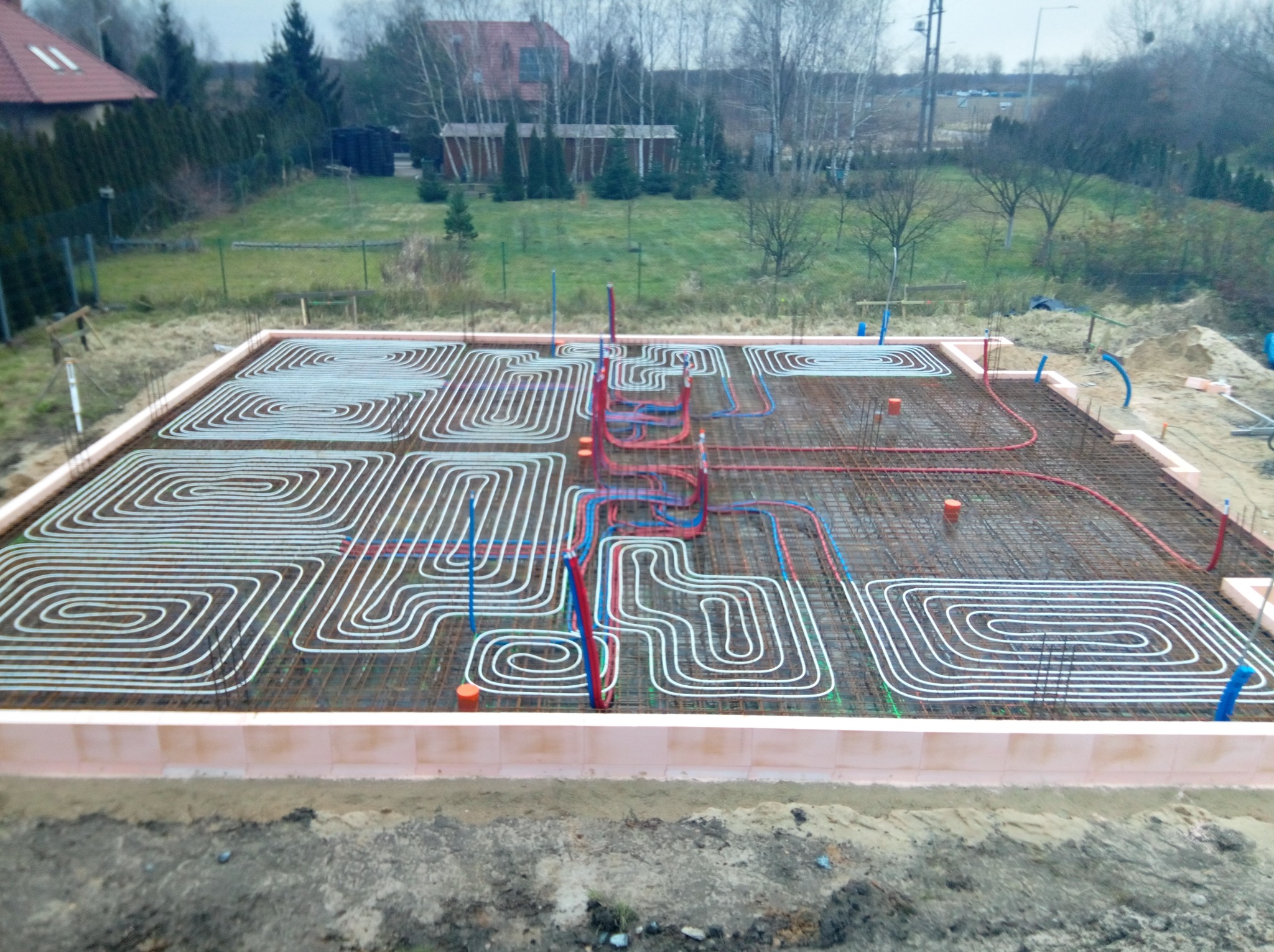 Przygotowany fundament z izolacją obwodową i rozłożonymi rurami ogrzewania podłogowego w kształcie meandrów, widoczne zbrojenie i pionowe rury instalacyjne.