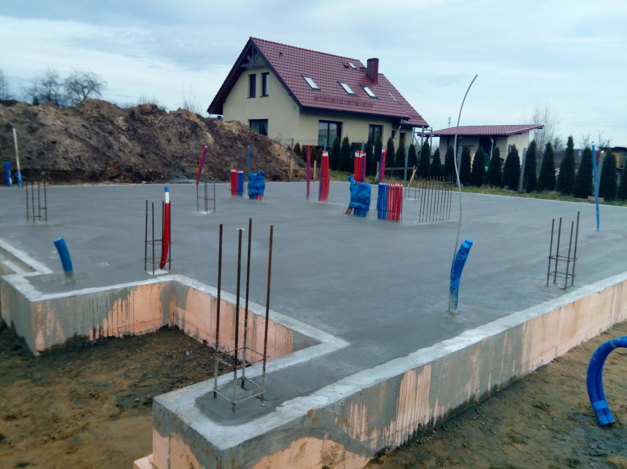 Zalany betonowy fundament z wystającymi prętami zbrojeniowymi i rurami instalacyjnymi, widok na dom z czerwonym dachem i stertę ziemi w tle, pochmurne niebo.