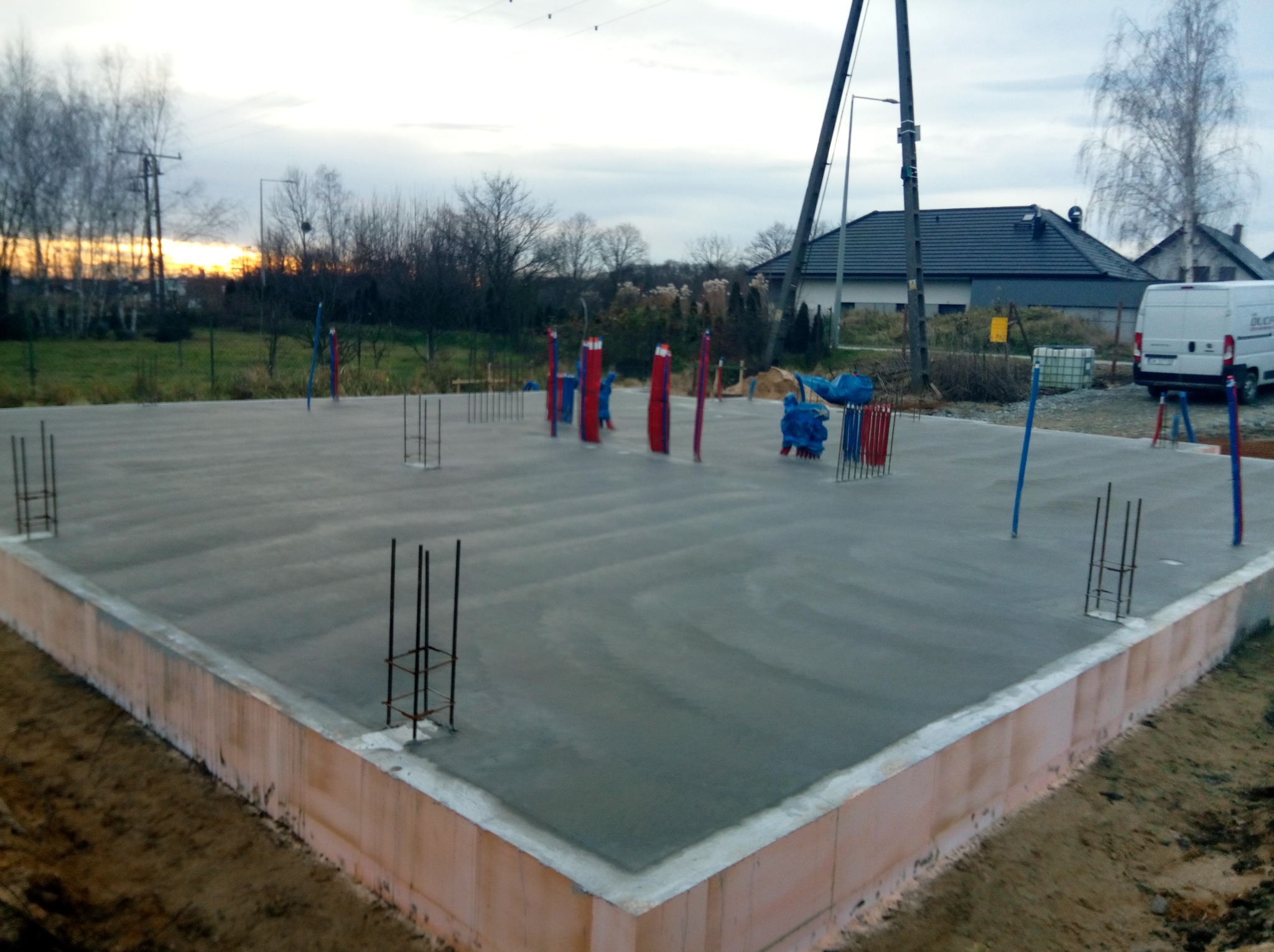 Wylana płyta fundamentowa z wystającymi prętami zbrojeniowymi, zabezpieczona styropianem, na tle zachodzącego słońca i zabudowy jednorodzinnej.