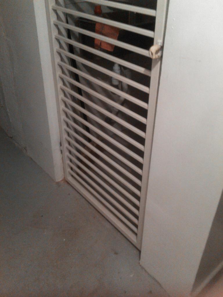 Metalowa krata z poziomymi szczeblami, zamontowana w ościeżnicy, widoczna z bliska, z fragmentem ściany w tle.