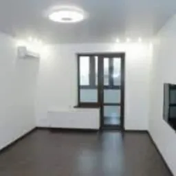 Puste, jasne pomieszczenie z parkietem w odcieniach brązu, białymi ścianami, klimatyzacją, oknem balkonowym z ciemną ramą i zamontowanym telewizorem na ścianie.