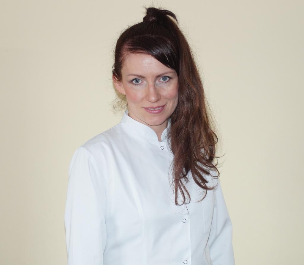 Portret kobiety w białym uniformie medycznym, z włosami upiętymi w wysoki kucyk, na jednolitym, kremowym tle.