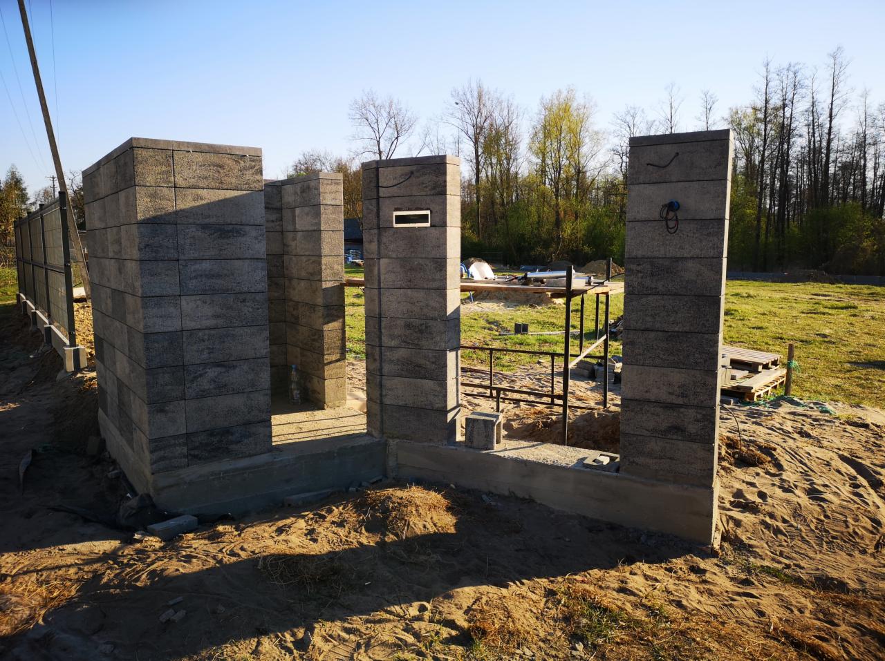 Wznoszone z szarej kostki filary ogrodzenia na fundamencie, widoczne narzędzia i rusztowanie w tle, trawa i drzewa w oddali.