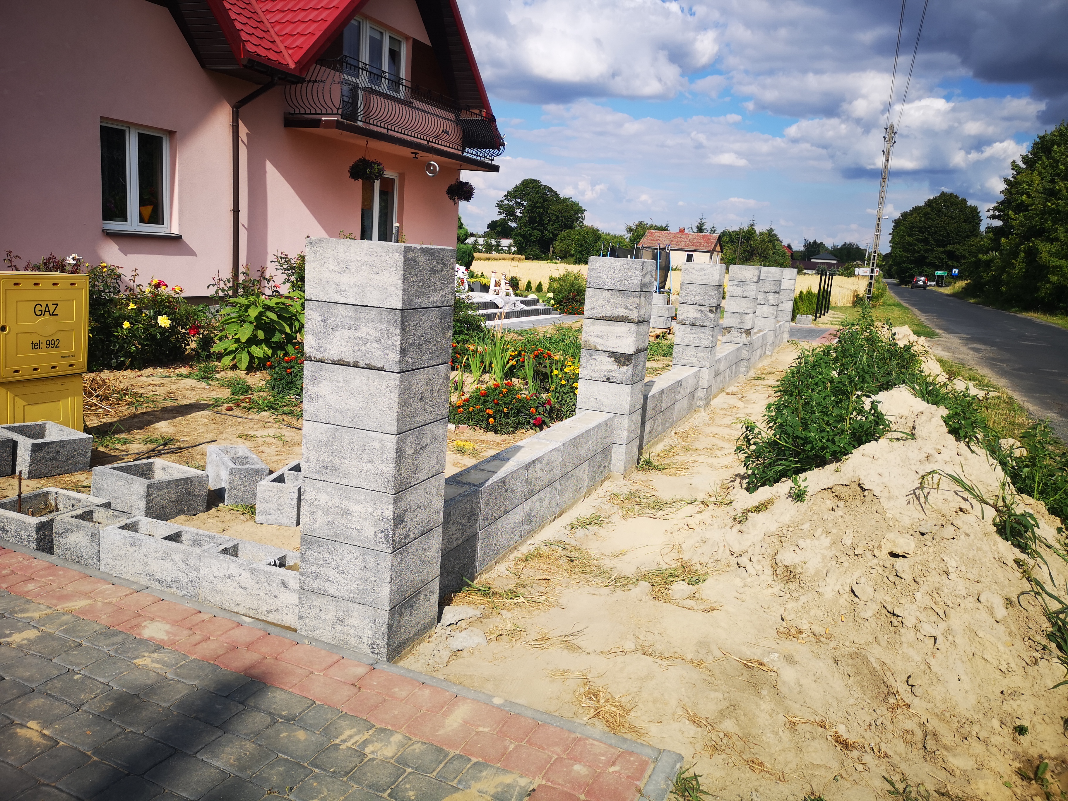 Budowa ogrodzenia z szarych betonowych bloczków modułowych, widoczne słupki i fragment muru, przygotowany teren, kostka brukowa na podjeździe, w tle dom z czerwonym dachem i skrzynka gazowa.
