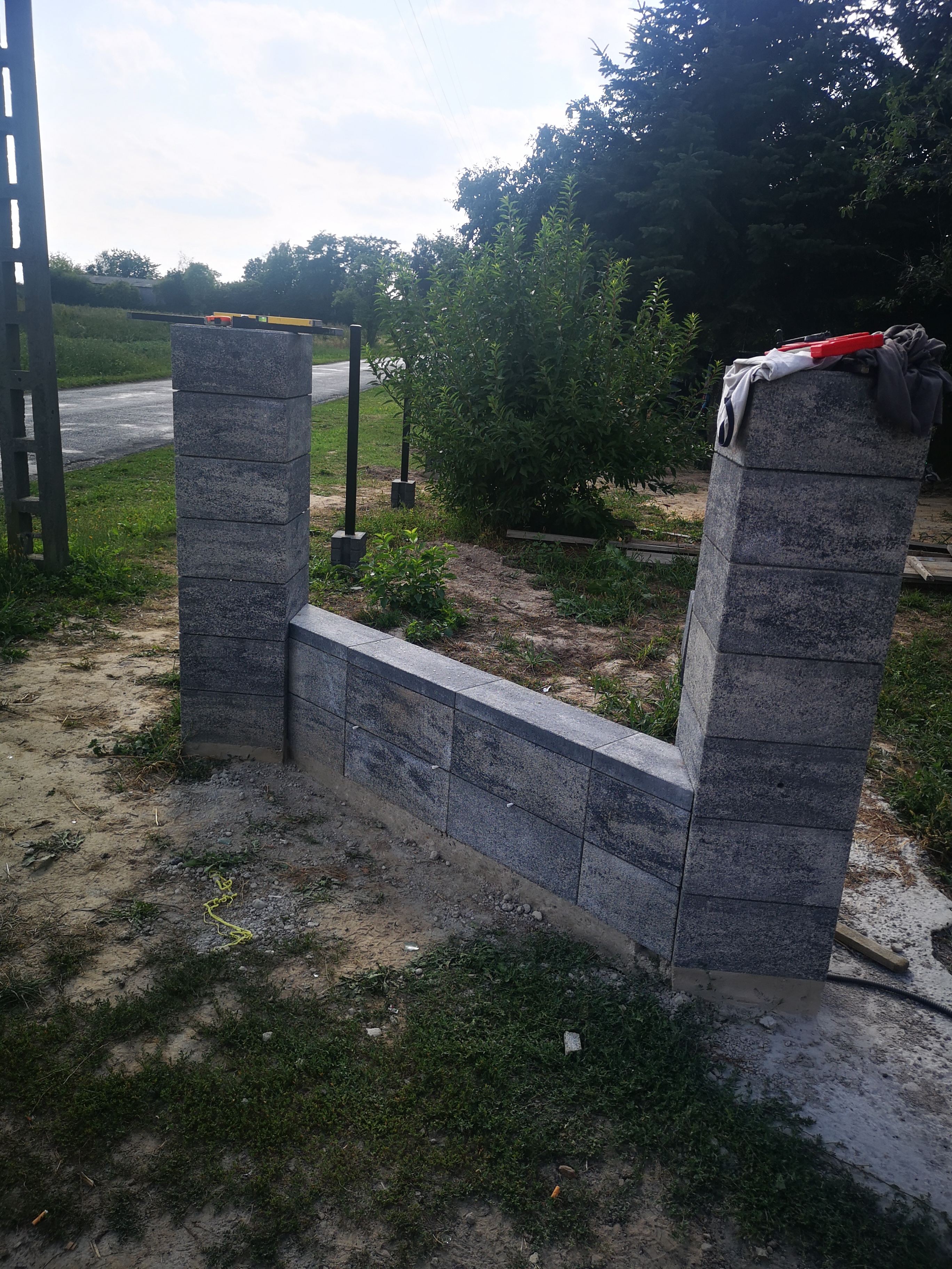 Fragment ogrodzenia z szarej kostki, w trakcie montażu, z widocznymi elementami konstrukcyjnymi i narzędziami. Tło stanowią drzewa i trawa.