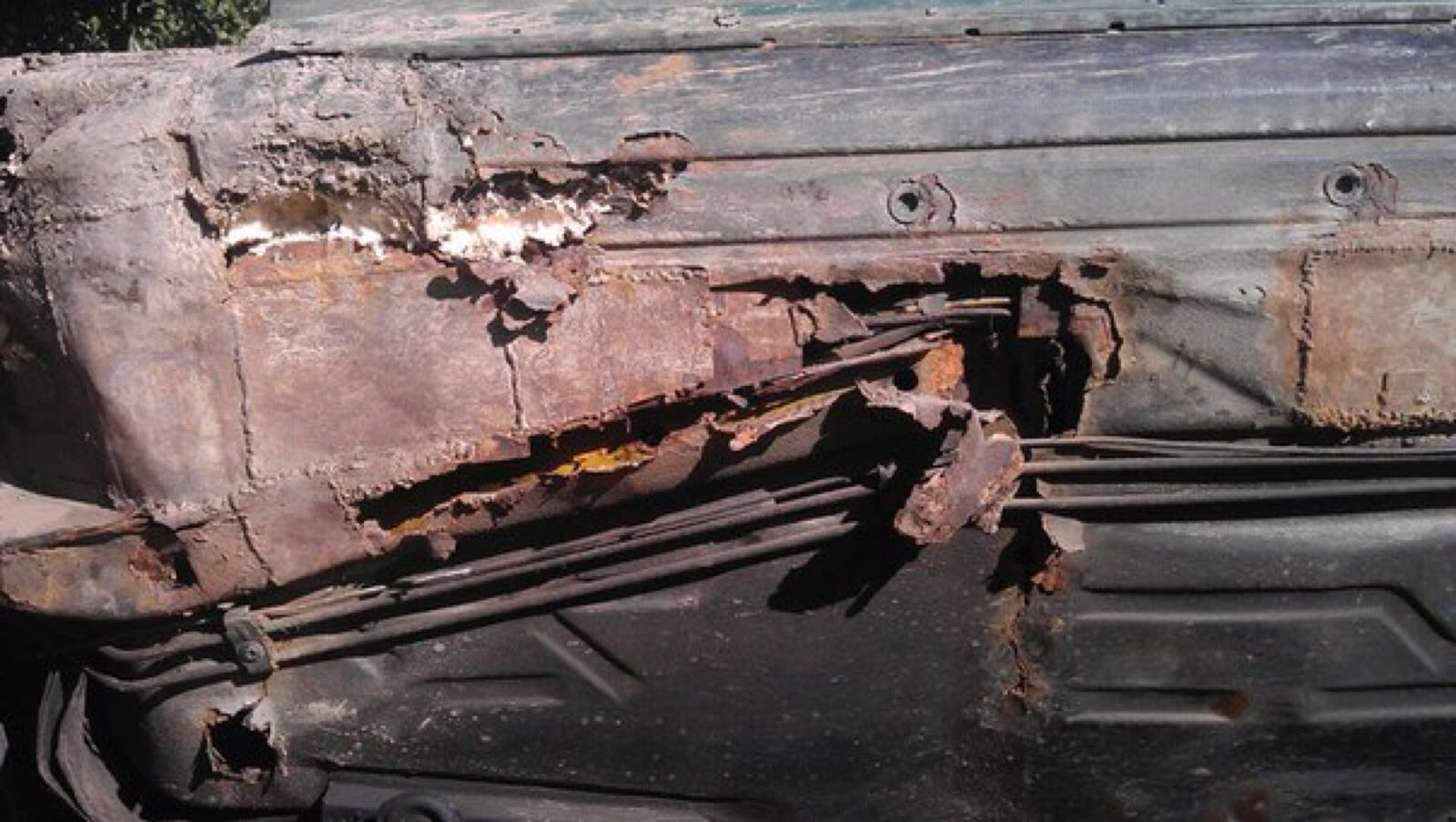 Silnie skorodowany fragment karoserii samochodu, z widocznymi dziurami, rdzawymi nalotami i uszkodzeniami powłoki lakierniczej, ukazujący przewody i wewnętrzną strukturę pojazdu.