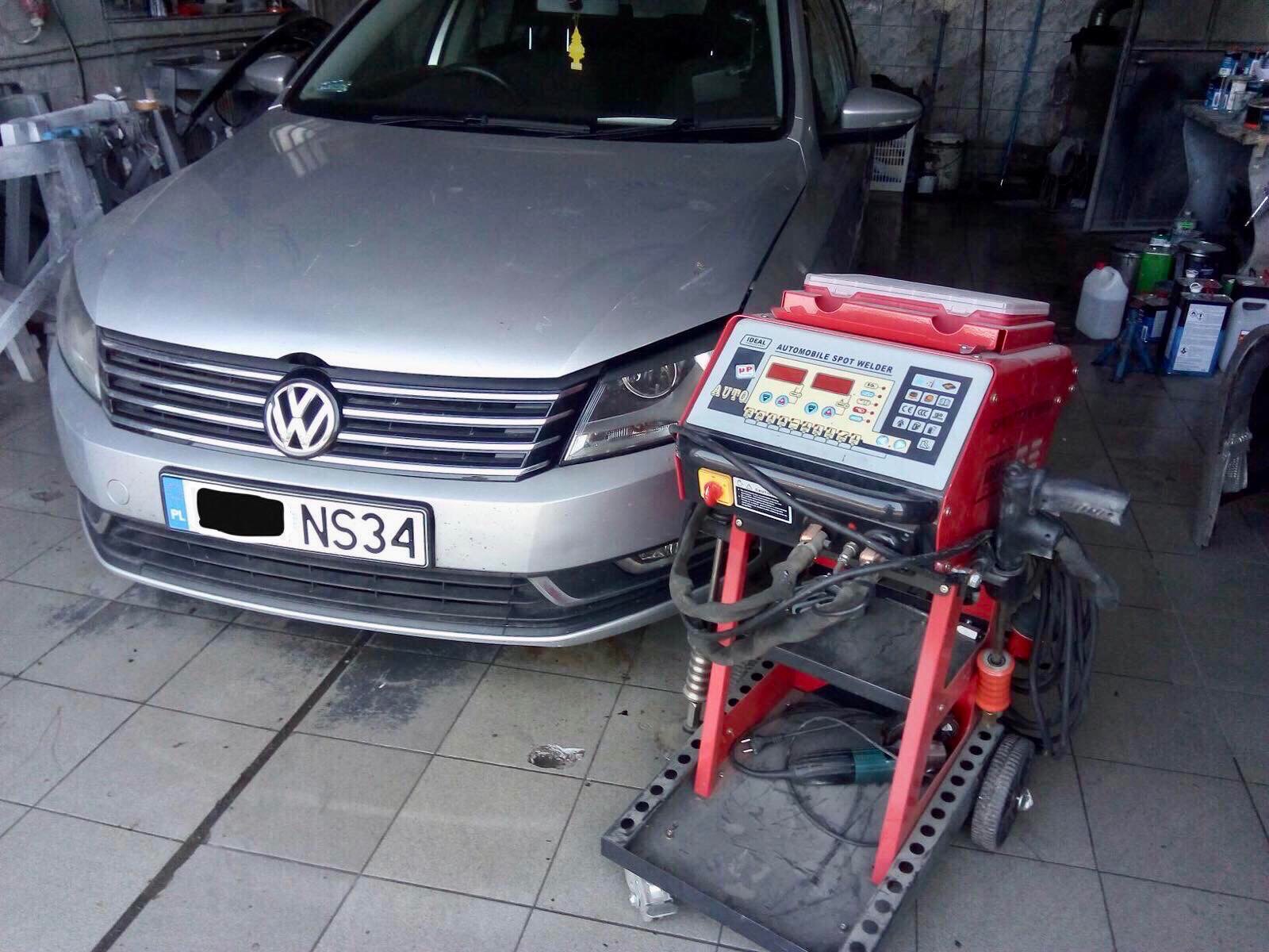 Srebrny Volkswagen Passat przed warsztatem, obok czerwone urządzenie do zgrzewania punktowego z widocznym panelem sterowania i przewodami.