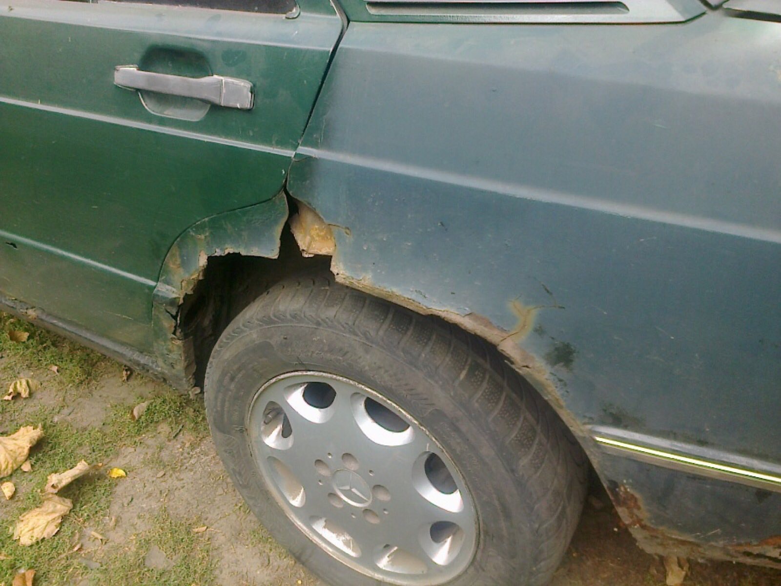 Zardzewiały, zielony samochód z uszkodzonym nadkolem i widoczną korozją wokół opony, na tle ziemi i opadłych liści.