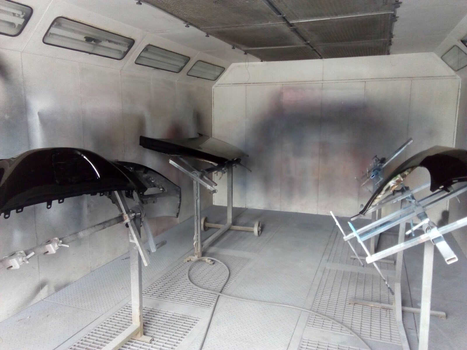 Elementy karoserii samochodowej, błotniki i maska, po lakierowaniu na wysoki połysk, zawieszone w kabinie lakierniczej na metalowych stojakach.