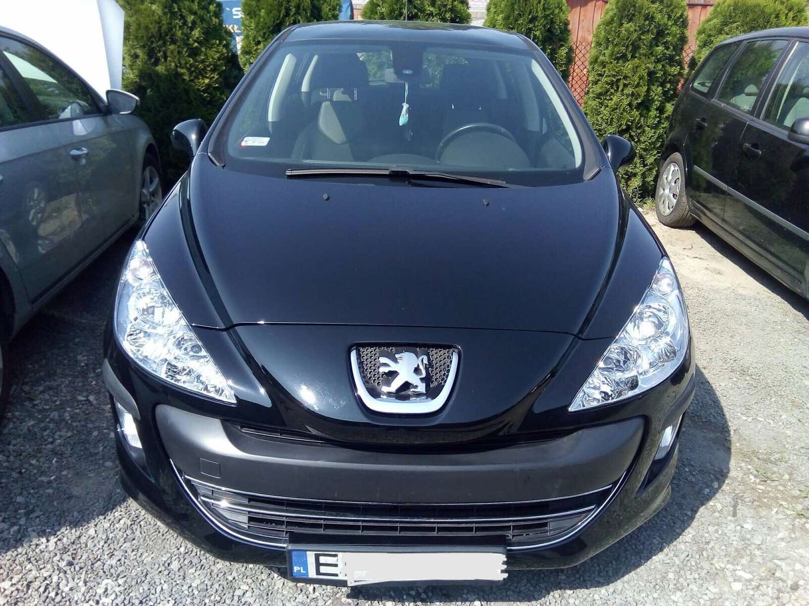 Błyszczący czarny samochód osobowy marki Peugeot zaparkowany na żwirowym podłożu, widok z przodu na maskę, reflektory i logo. W tle żywopłot i inne pojazdy.