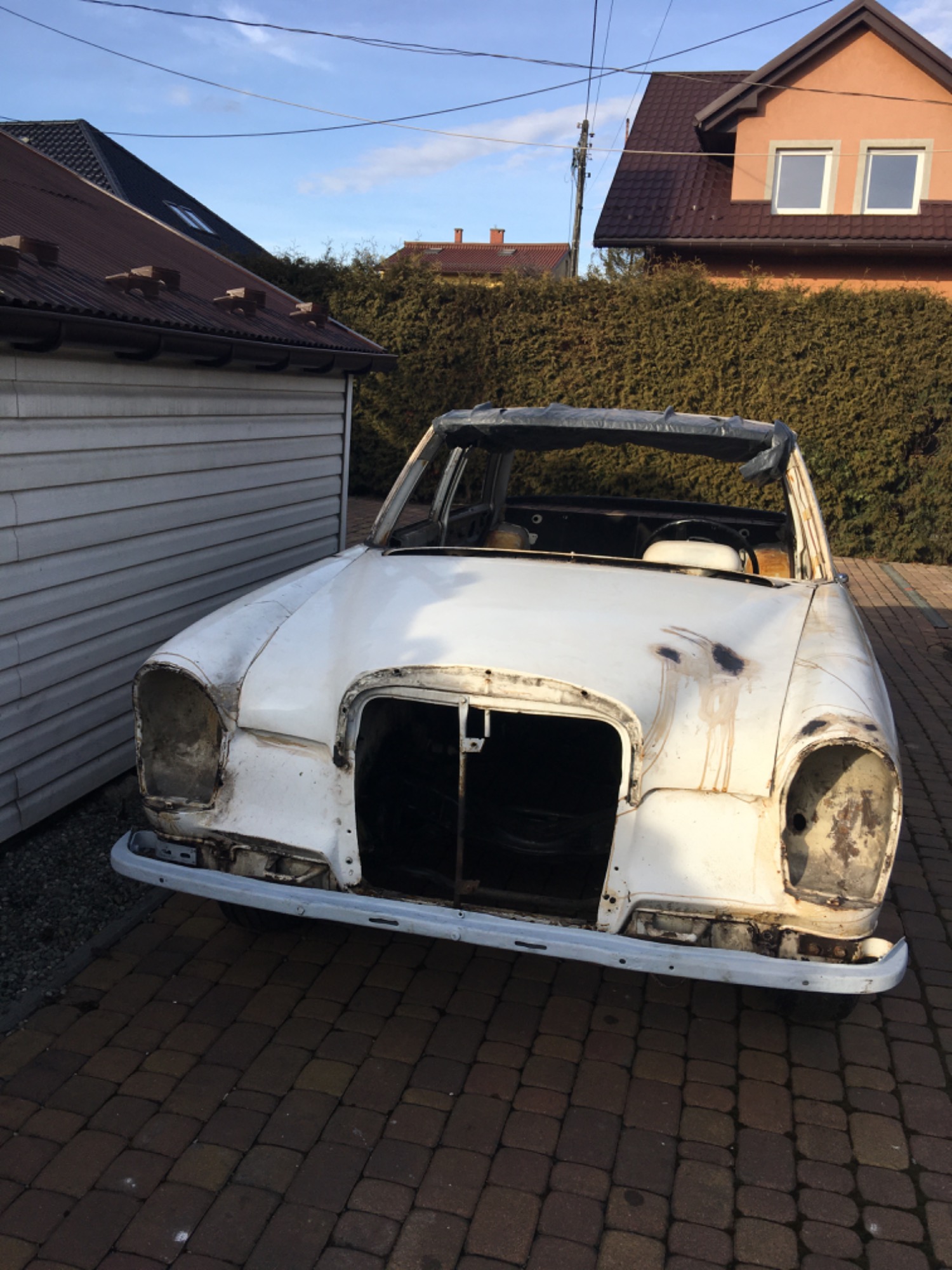 Zdjęcie białego, zdekompletowanego samochodu marki Mercedes, bez reflektorów i z widocznymi śladami korozji, stojącego na brukowanym podjeździe przed budynkami i żywopłotem.