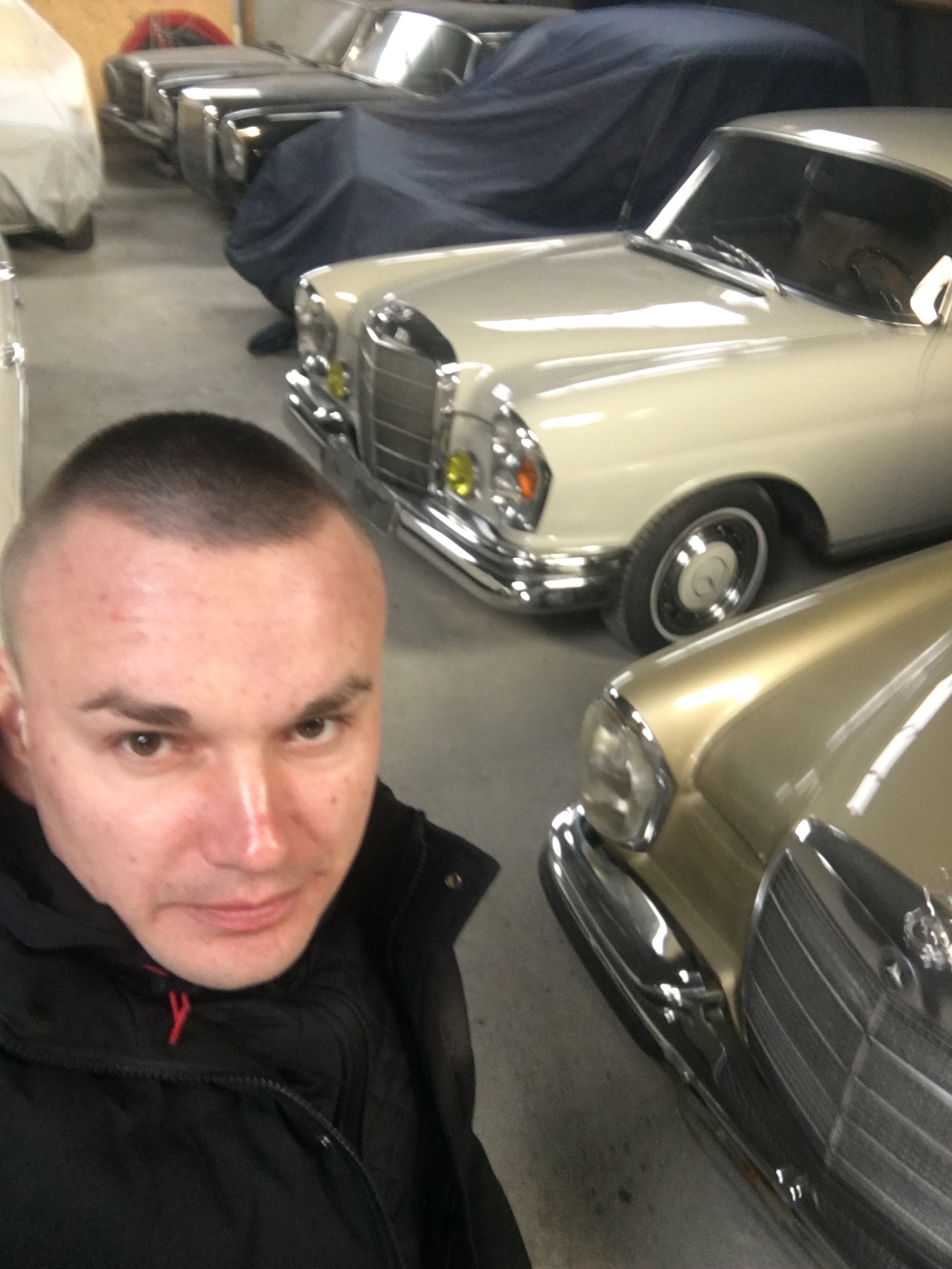 Selfie mężczyzny na tle klasycznych Mercedesów w garażu, jeden przykryty plandeką, widoczne detale chromowanych elementów i żółte reflektory.
