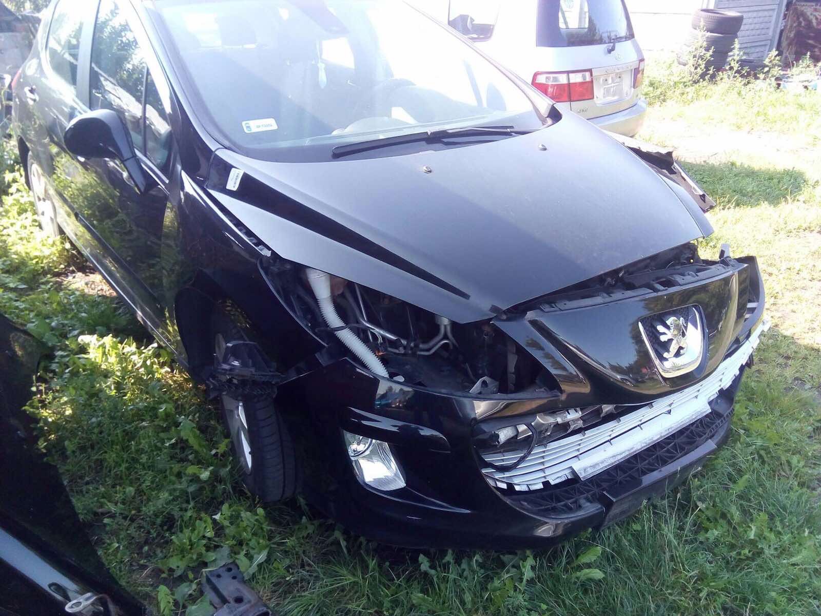 Uszkodzony przód czarnego samochodu osobowego marki Peugeot, widoczne uszkodzenia karoserii i brakujące elementy, auto stoi na trawie.